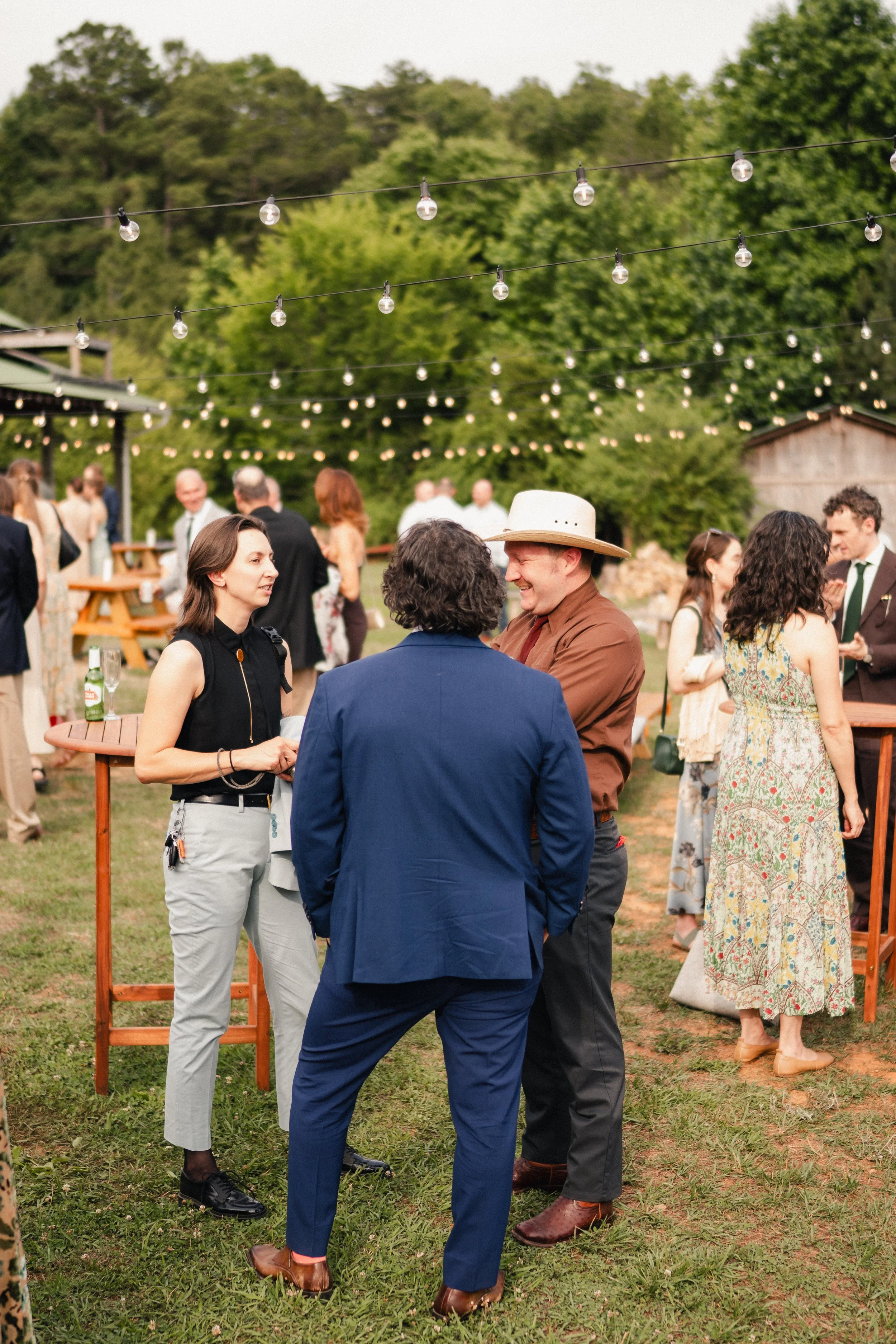 People socializing at an outdoor gathering under string lights, with trees and a barn in the background.