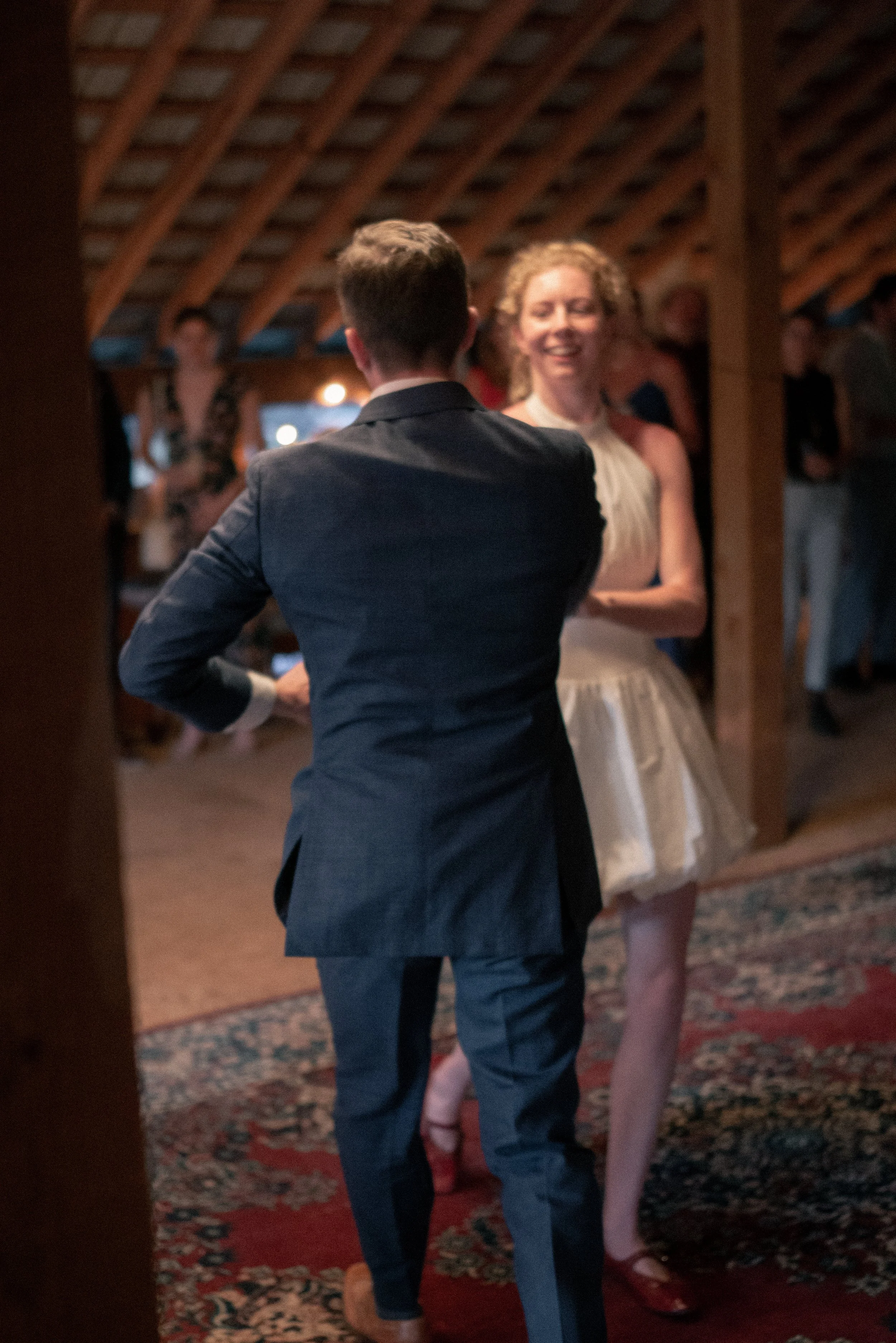 A man in a dark suit with short hair, seen from the back, facing a woman in a light-colored dress with curly hair, both smiling and dancing in a dimly lit room with a patterned carpet and wooden ceiling.