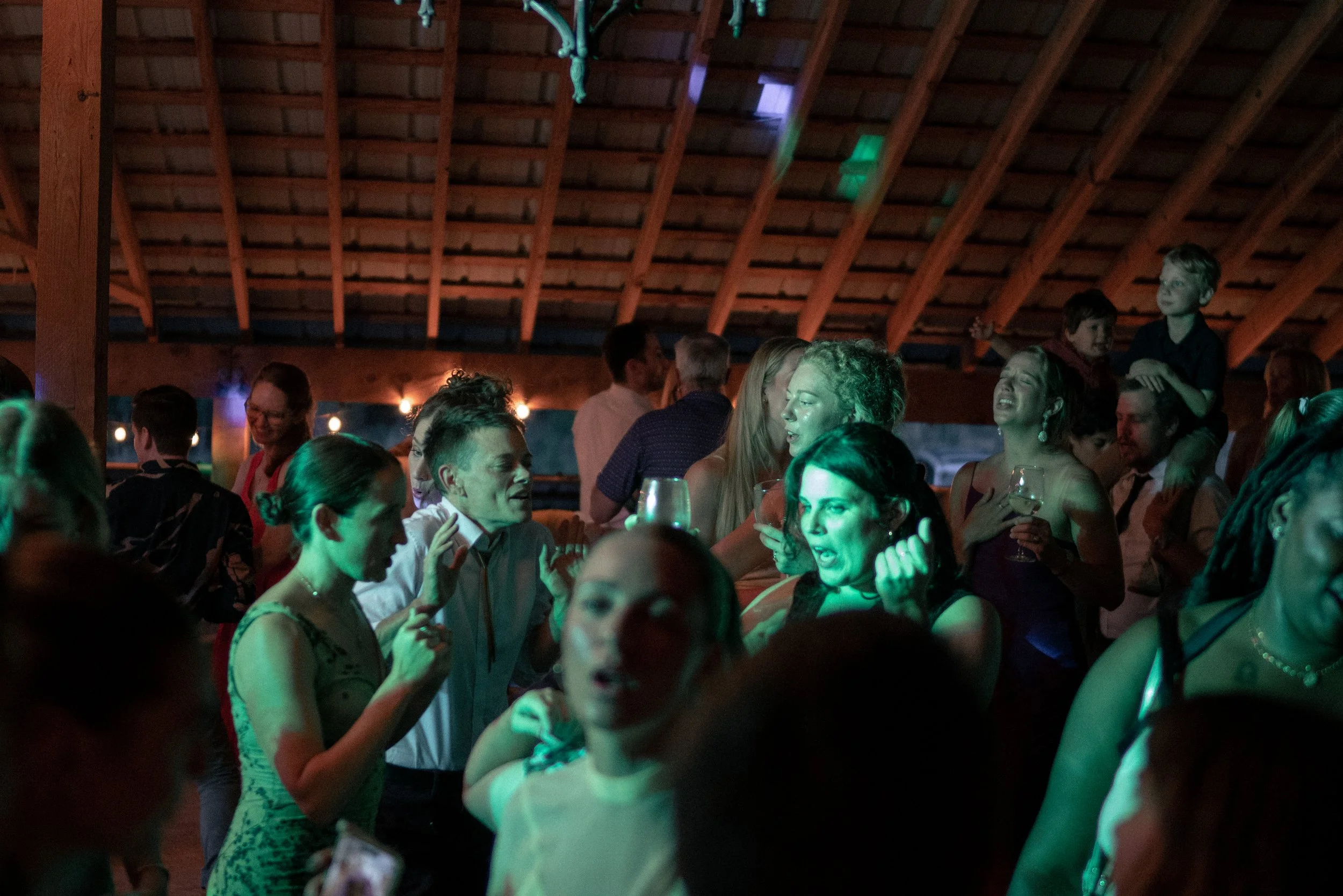 People gathered at an indoor event, dancing and socializing, with a wooden ceiling and colorful lighting.