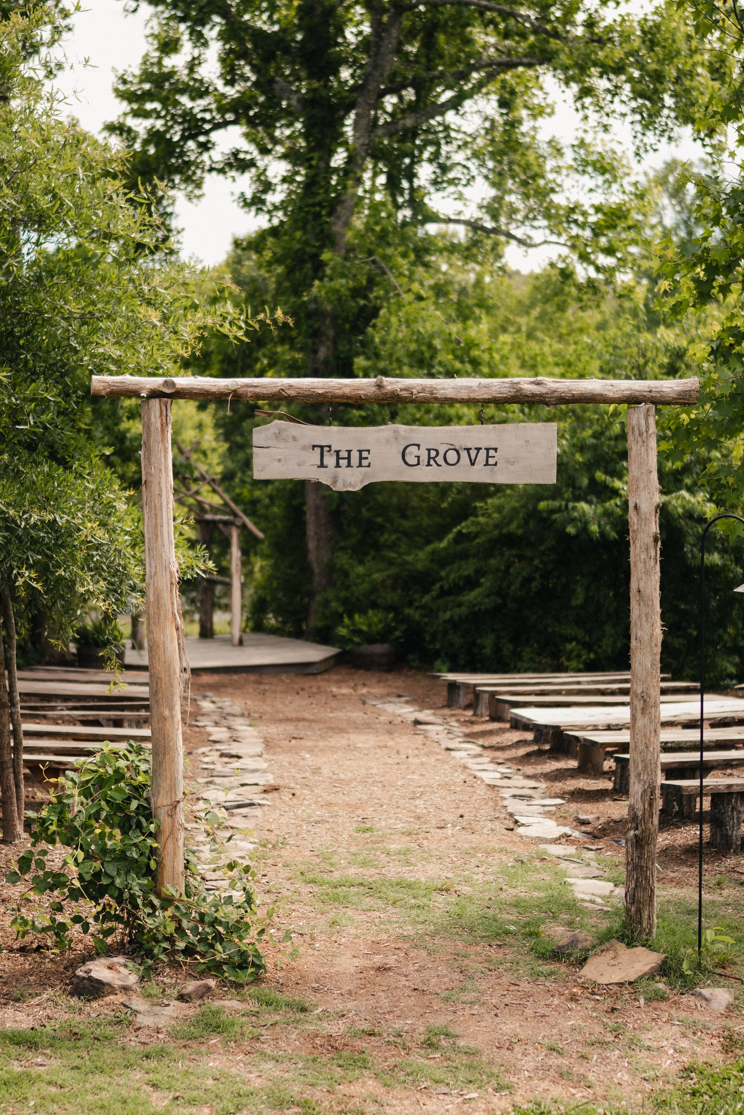 A wooden sign reading 'The Grove' hangs above a dirt pathway in a lush green outdoor setting, leading to a small stage area with wooden seating benches.