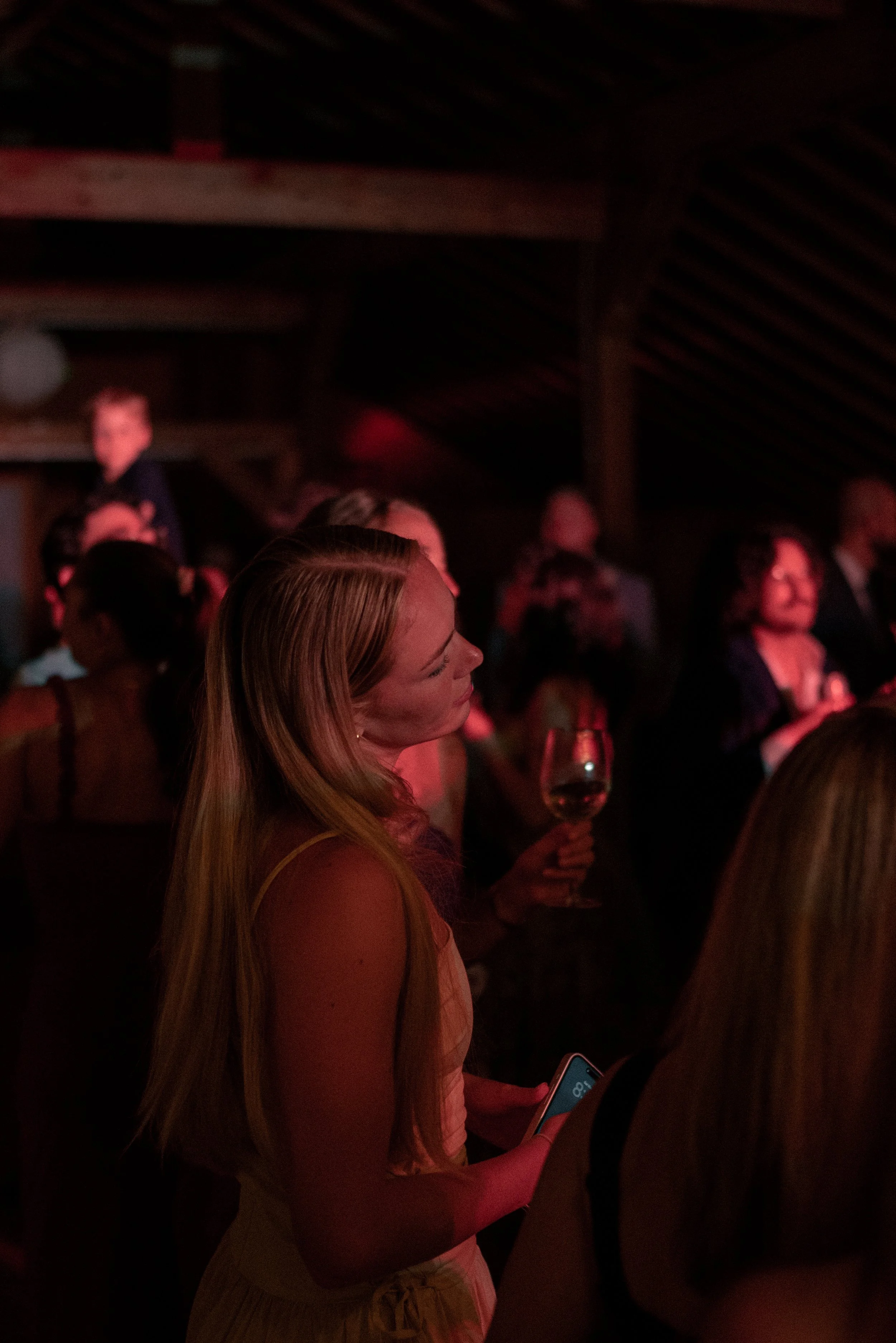 Young woman with long blonde hair holding a glass of wine, surrounded by other people in a dim indoor setting with wooden ceiling beams, warm red lighting, and a social atmosphere.