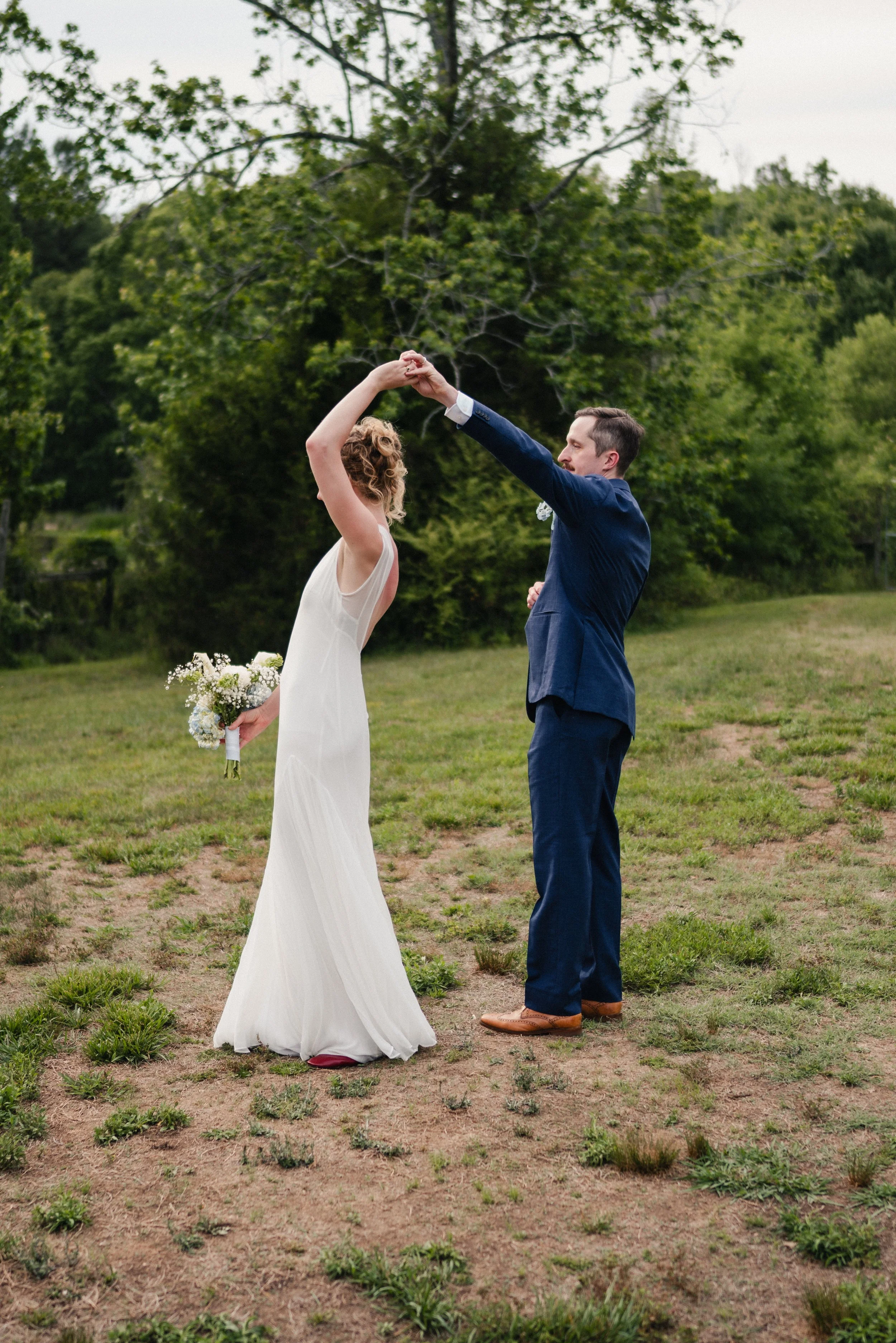 A bride and groom dancing outdoors on a grassy field with trees in the background.