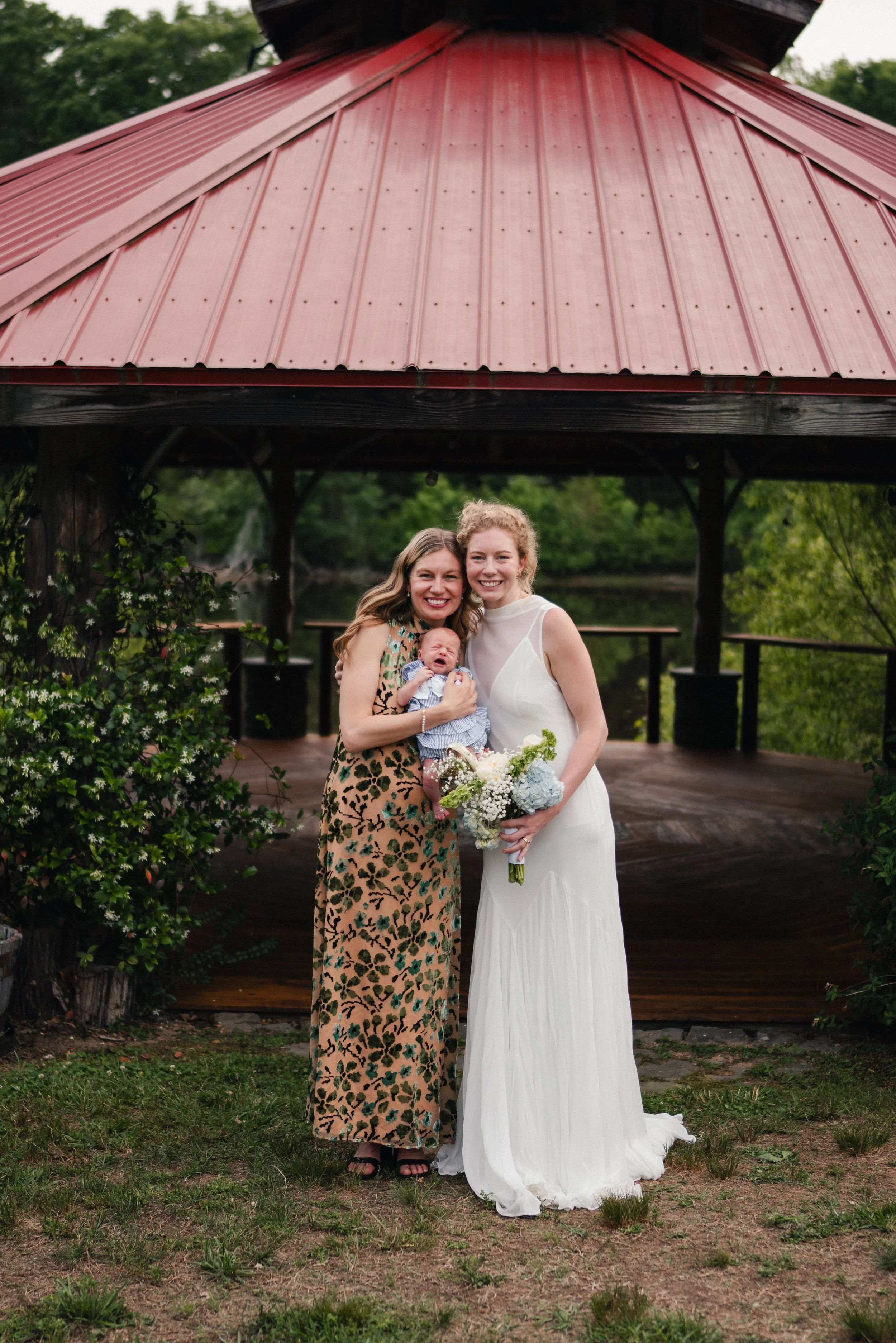 Two women, one in a white dress holding a bouquet and the other in a leopard print dress holding a crying baby, standing under a gazebo with a red metal roof in an outdoor park setting.