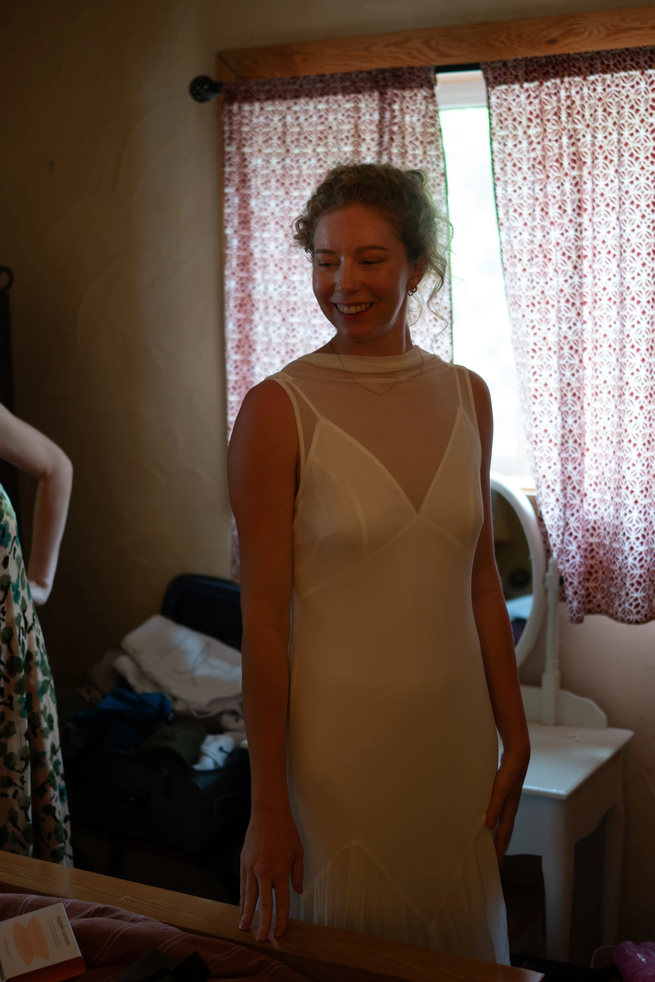 A woman wearing a white dress standing in a room with pink patterned curtains and a wooden headboard behind her, smiling.