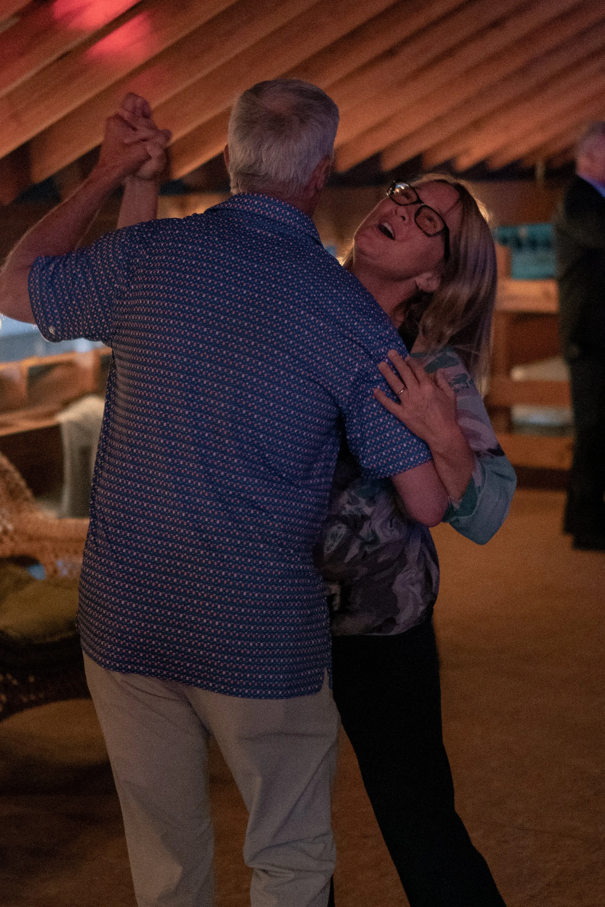 Older man and woman dancing together indoors, holding hands, with warm lighting and wooden ceiling in background.