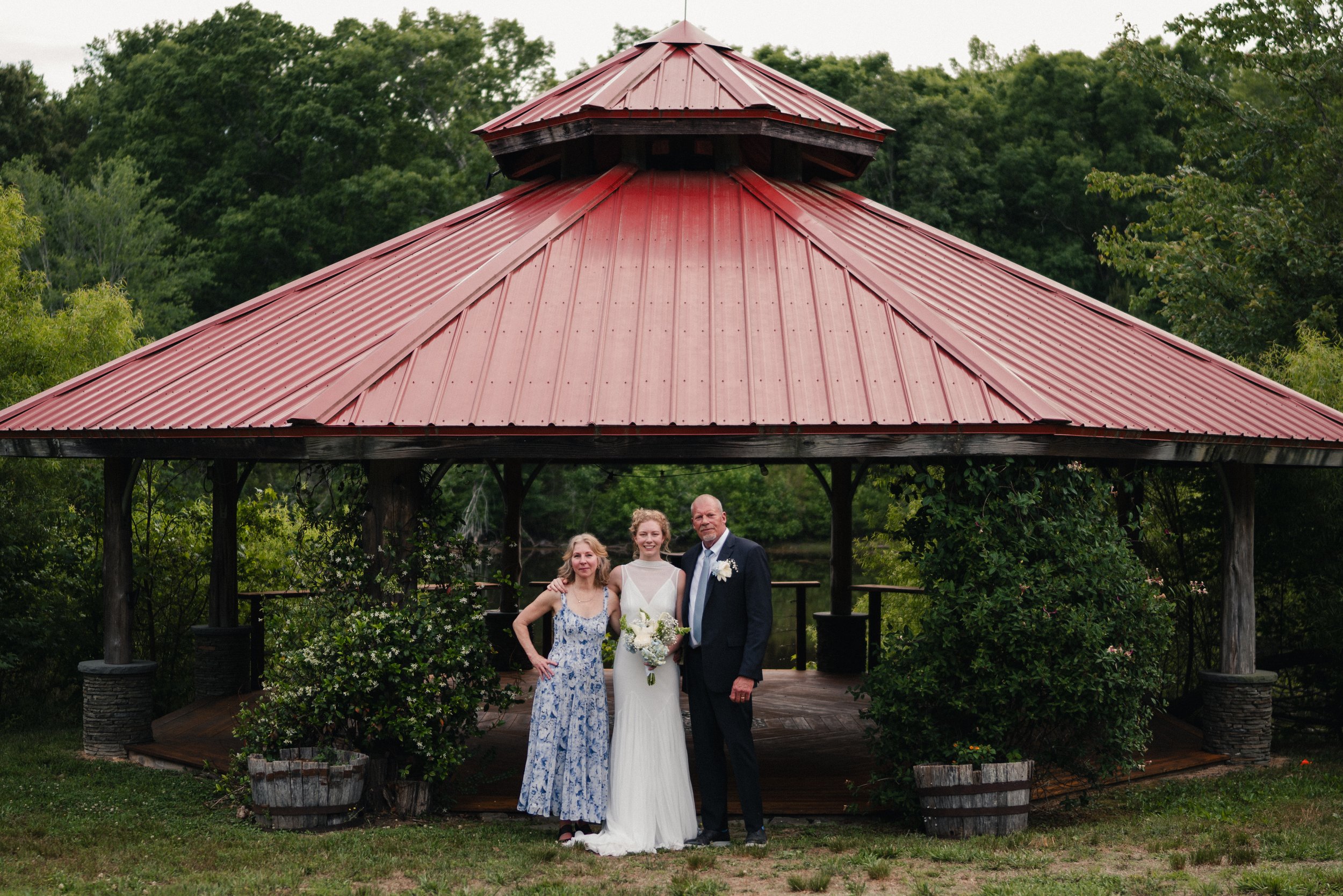 A group of four people, including a bride in a white wedding gown holding a bouquet, standing under a red-roofed pavilion surrounded by green trees and plants.