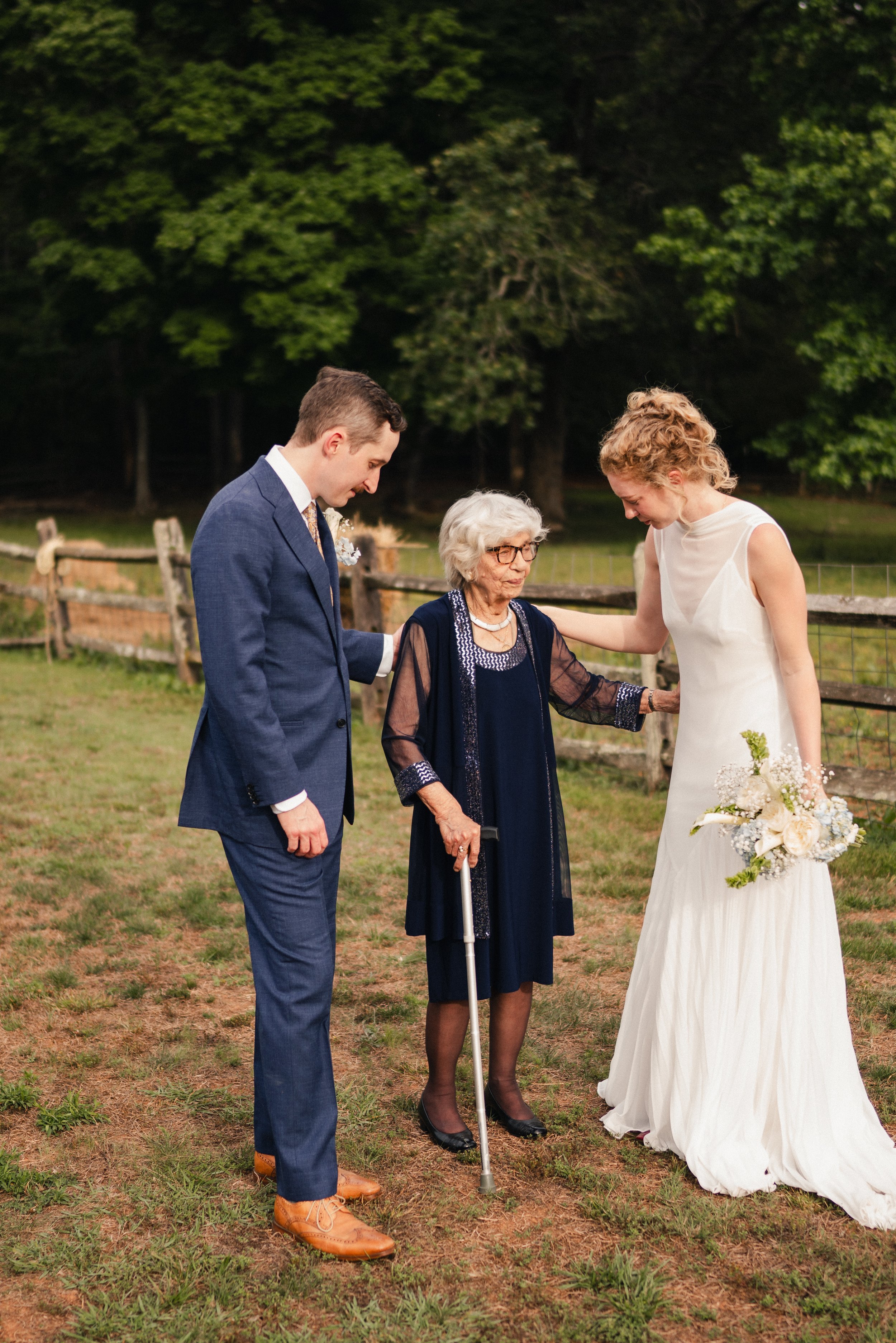 A wedding scene outdoors with a woman in a white dress holding a bouquet, two young adults in formal attire, and an elderly woman with a cane, all showing reverence or prayer.