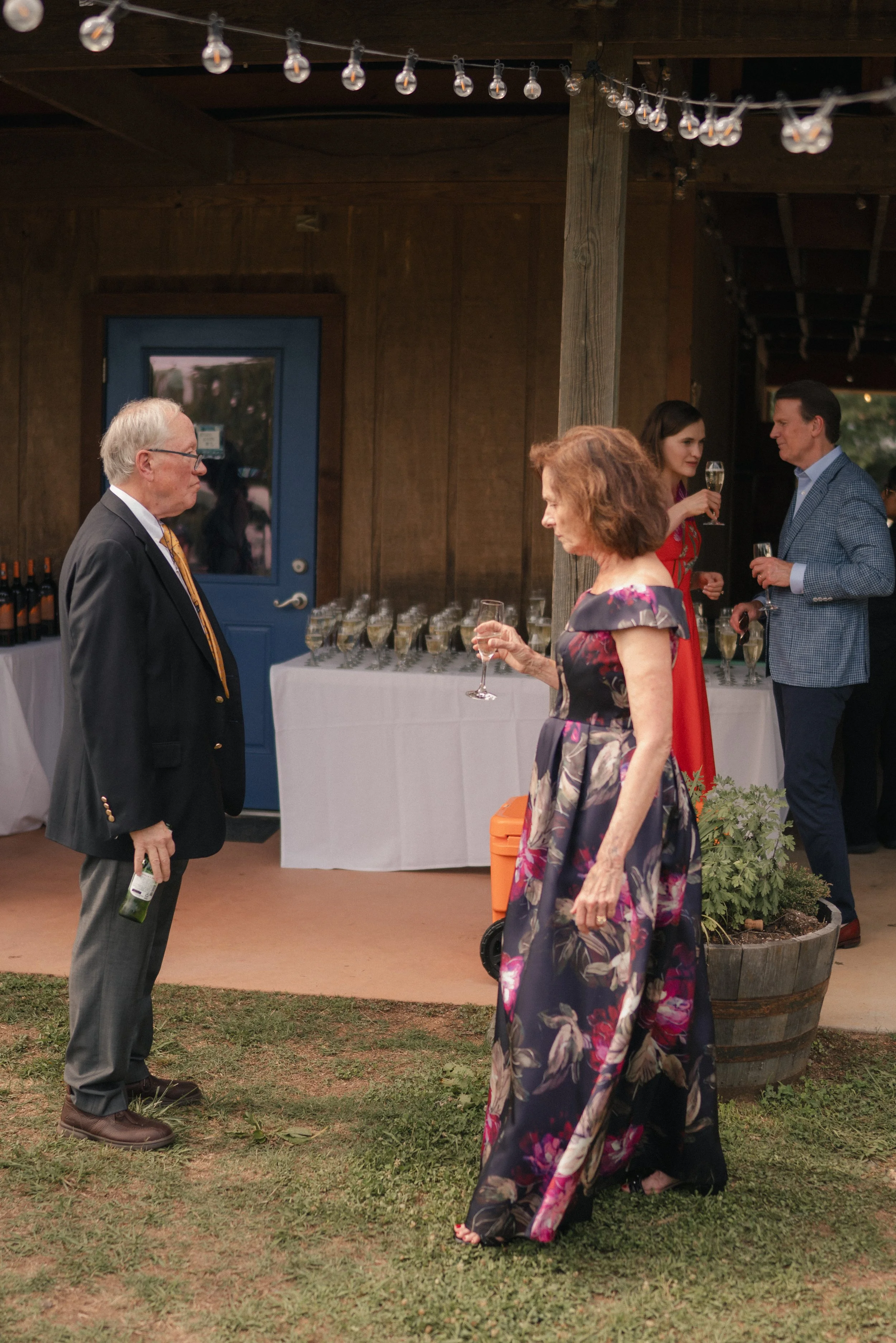 Four people in formal attire at an outdoor event, talking and holding glasses of champagne, with a table of champagne glasses and a decorated wooden shed in the background.