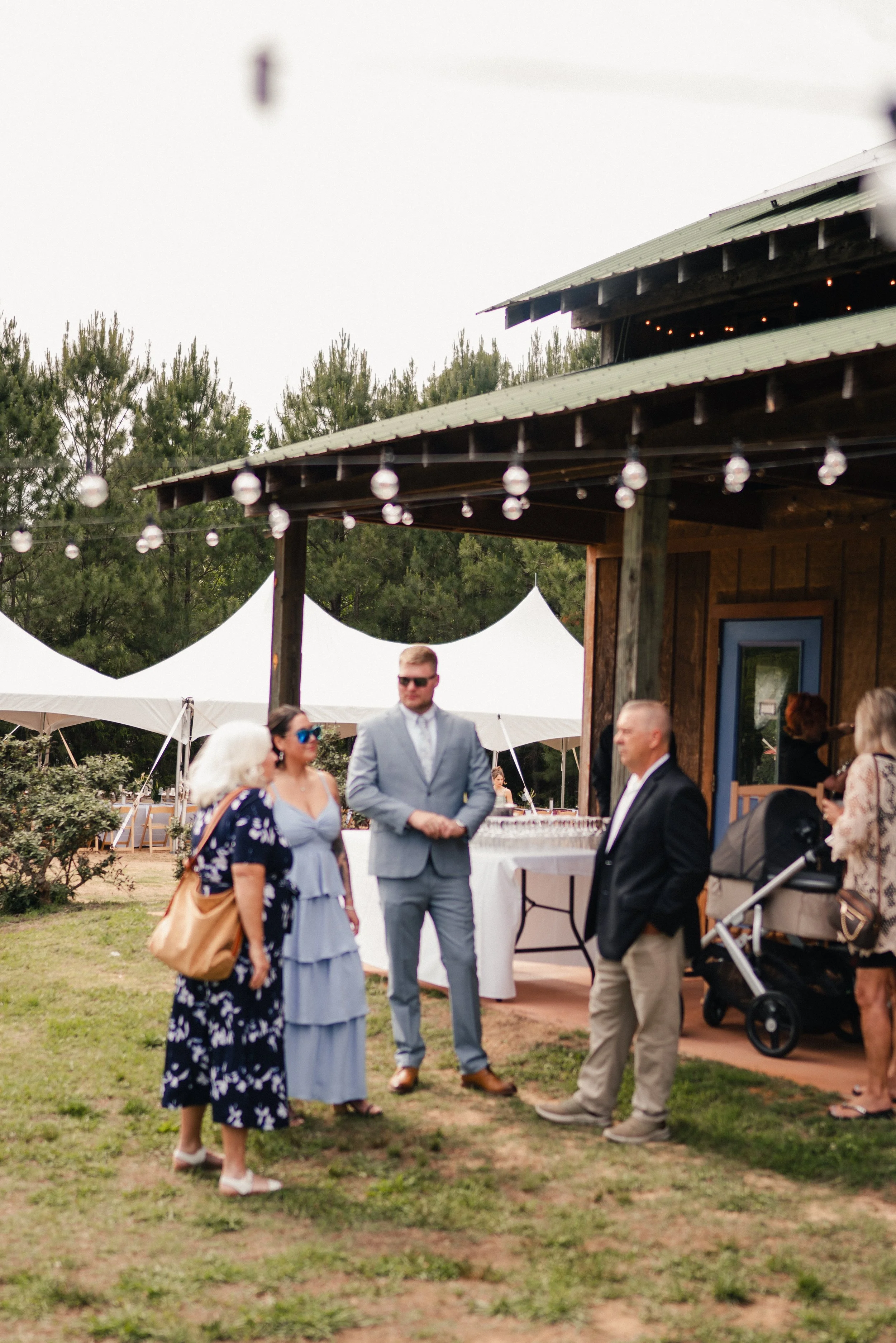 People dressed in formal and casual attire at an outdoor event with tents, string lights, and trees in the background.