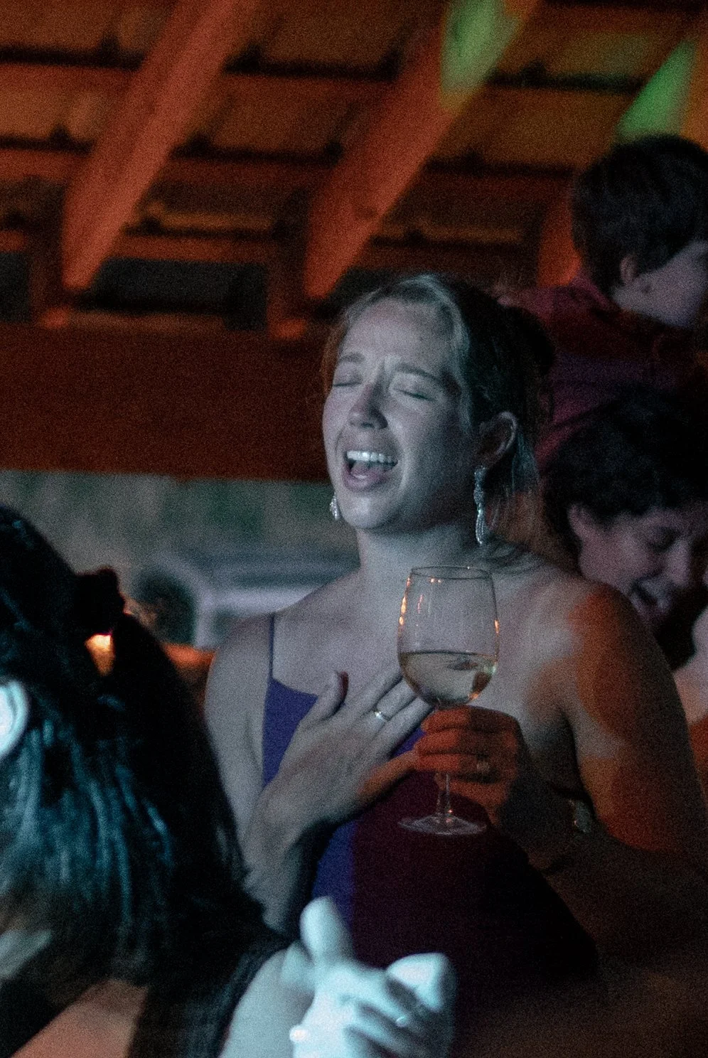 A woman in a purple dress at a party, singing and holding a glass of wine, with her hand on her chest and eyes closed, in a lively social setting.