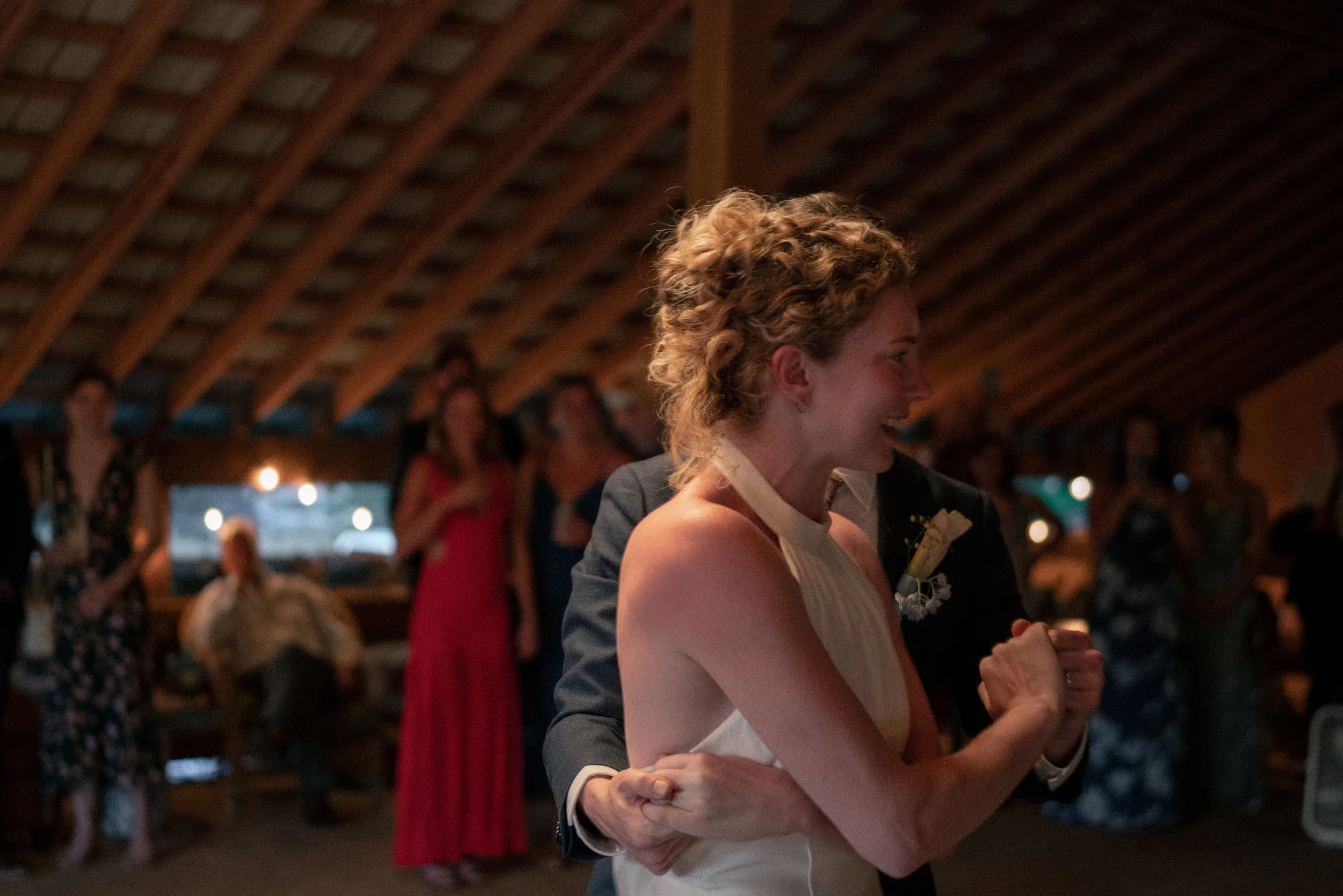 A woman with curly blonde hair wearing a white dress and a man in a dark suit and white shirt are dancing closely together, smiling, at a wedding reception inside a wooden venue with a vaulted ceiling. People are seen in the background, some standing