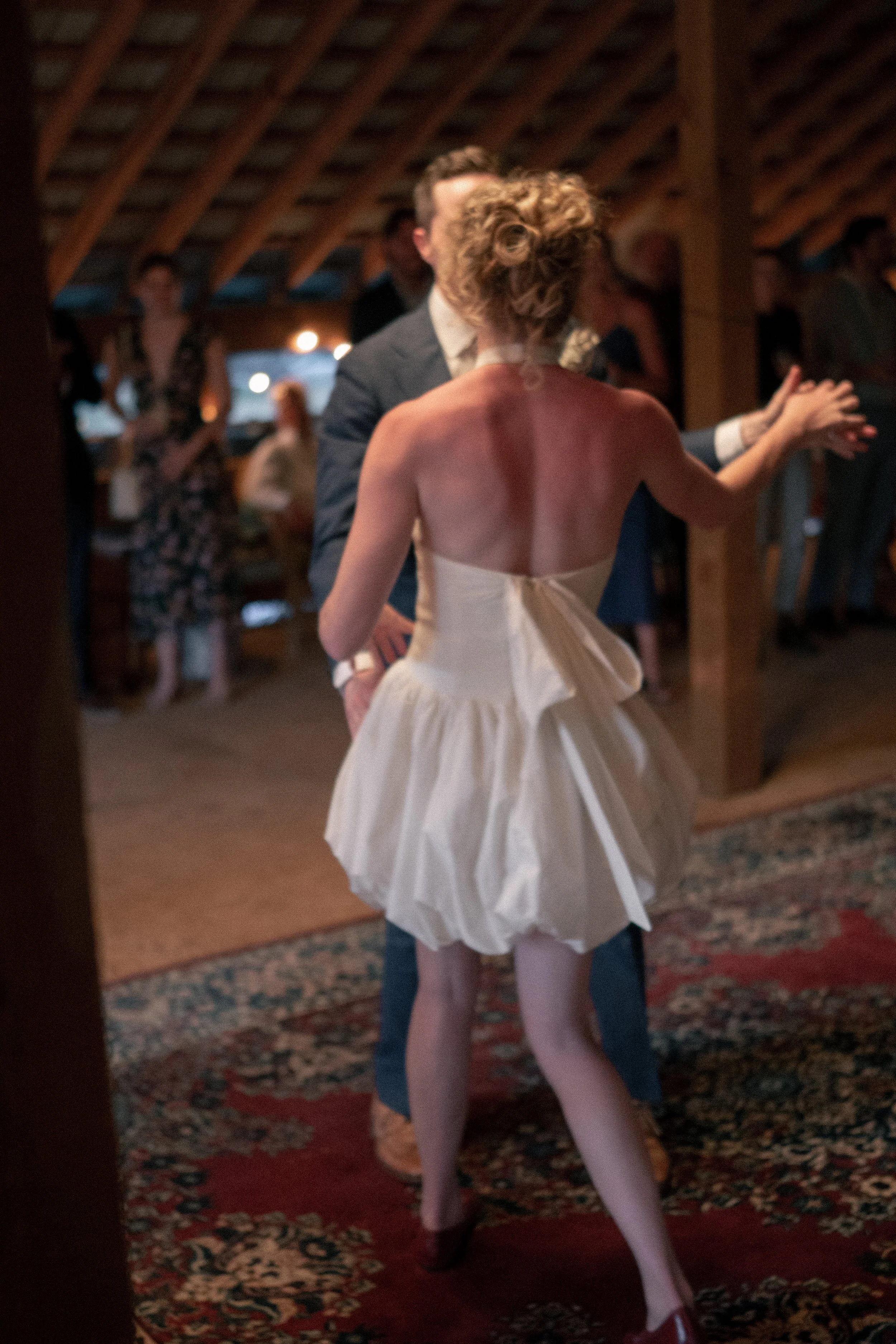 A woman in a white dress and pink tights dancing or twirling with a man in a suit during a party or celebration in a decorated indoor setting with people in the background.