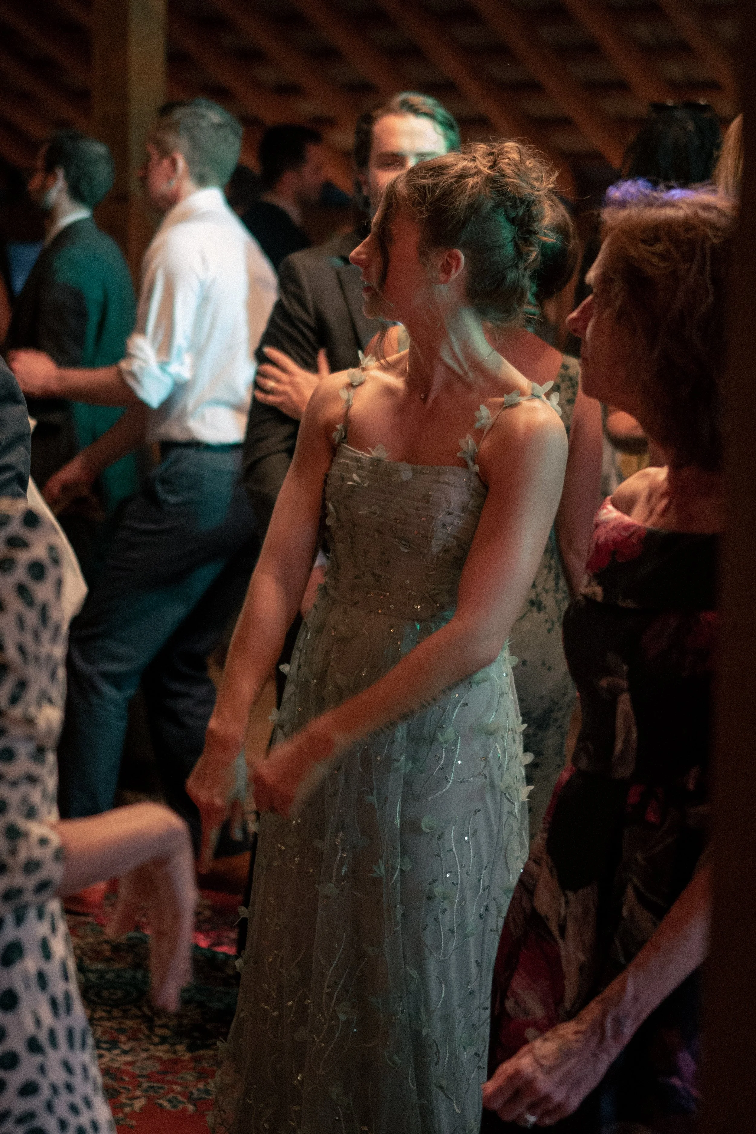 A woman in a strapless, silver gown with floral accents stands among guests at a formal event. She has her hair styled in an elegant updo and is engaged in conversation with a woman in a dark dress with a floral pattern. Other guests in formal attire