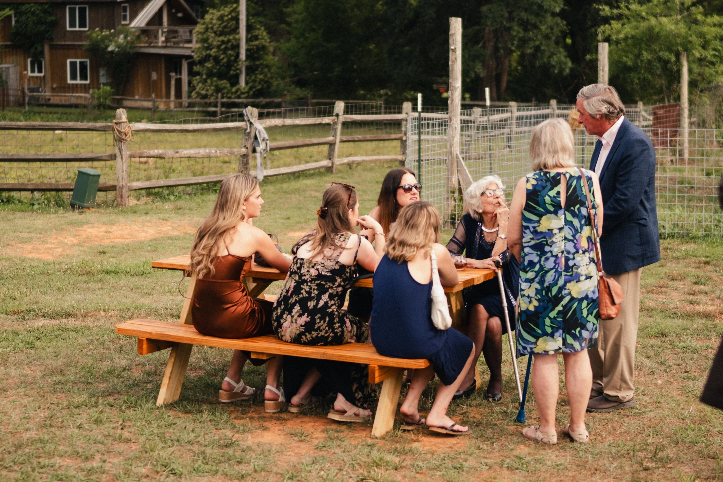 A group of women and an older man having a conversation outdoors in a park with wooden picnic tables and a fenced grassy area in the background.