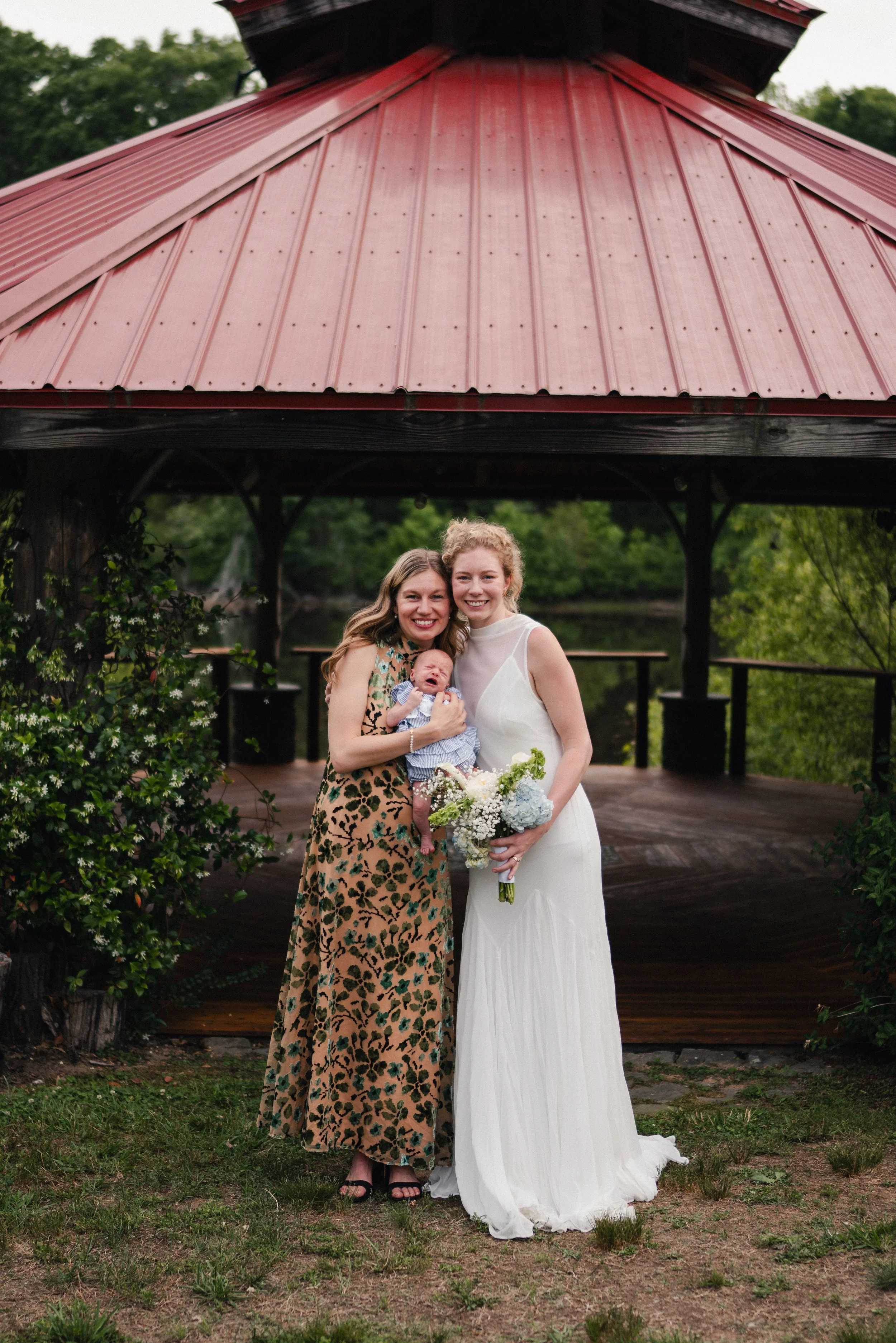 Two women, one in a white wedding dress holding a bouquet of flowers and a crying baby, stand close together under a pavilion with a red metal roof, greenery in the background.