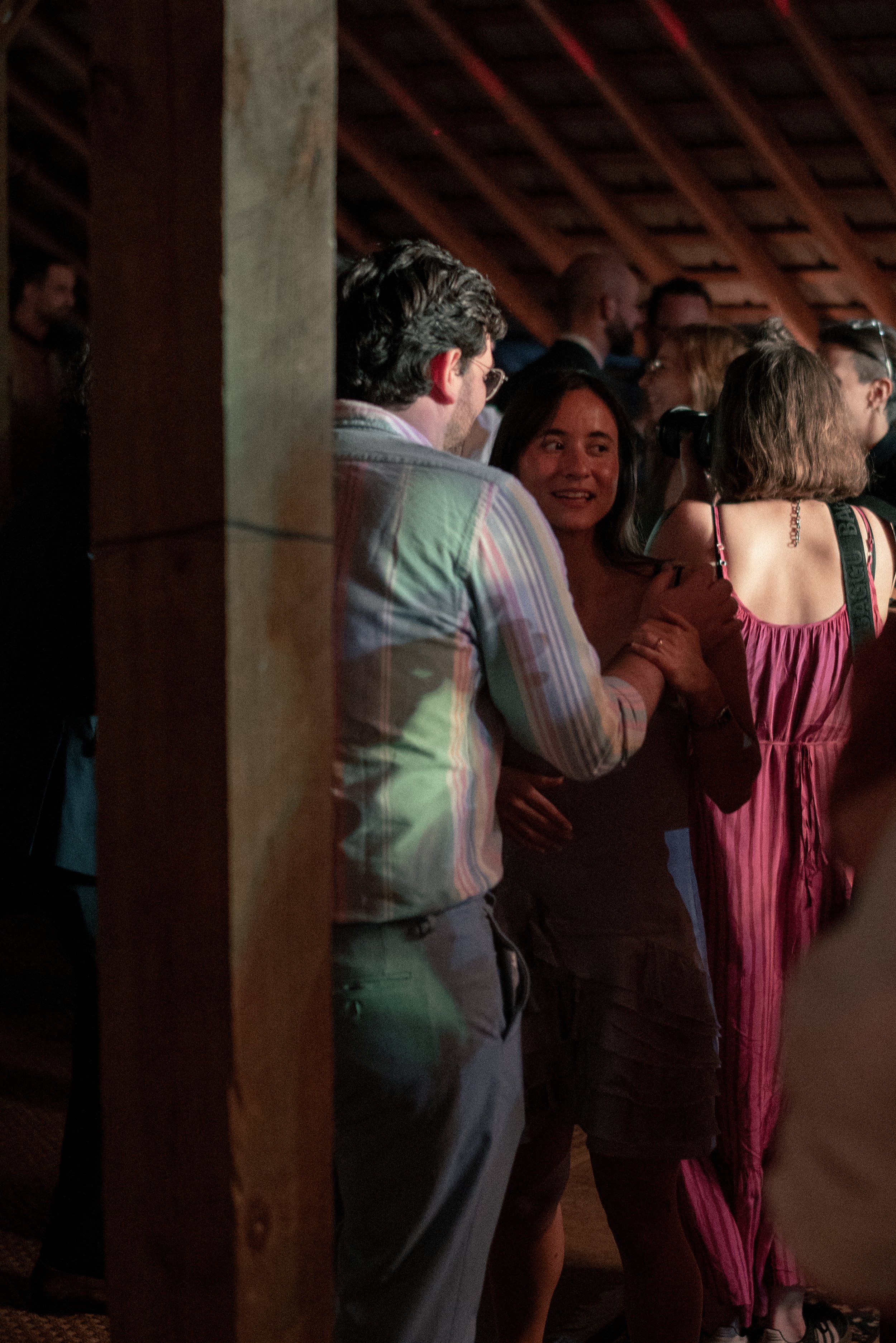 People gathered in a dimly lit room, engaged in conversation. The photo shows a man with dark, curly hair in a plaid shirt talking to a woman with long dark hair, wearing a brown top. Other individuals are visible in the background, some wearing glas