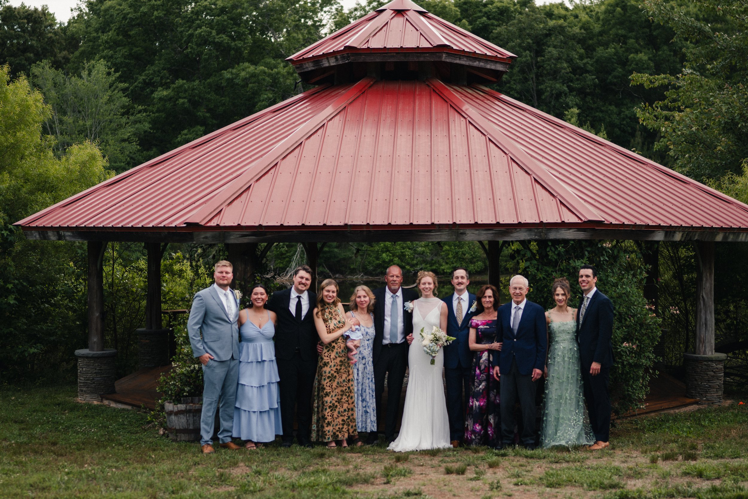 People gathered in wedding attire standing in front of a gazebo with a red roof, surrounded by greenery.