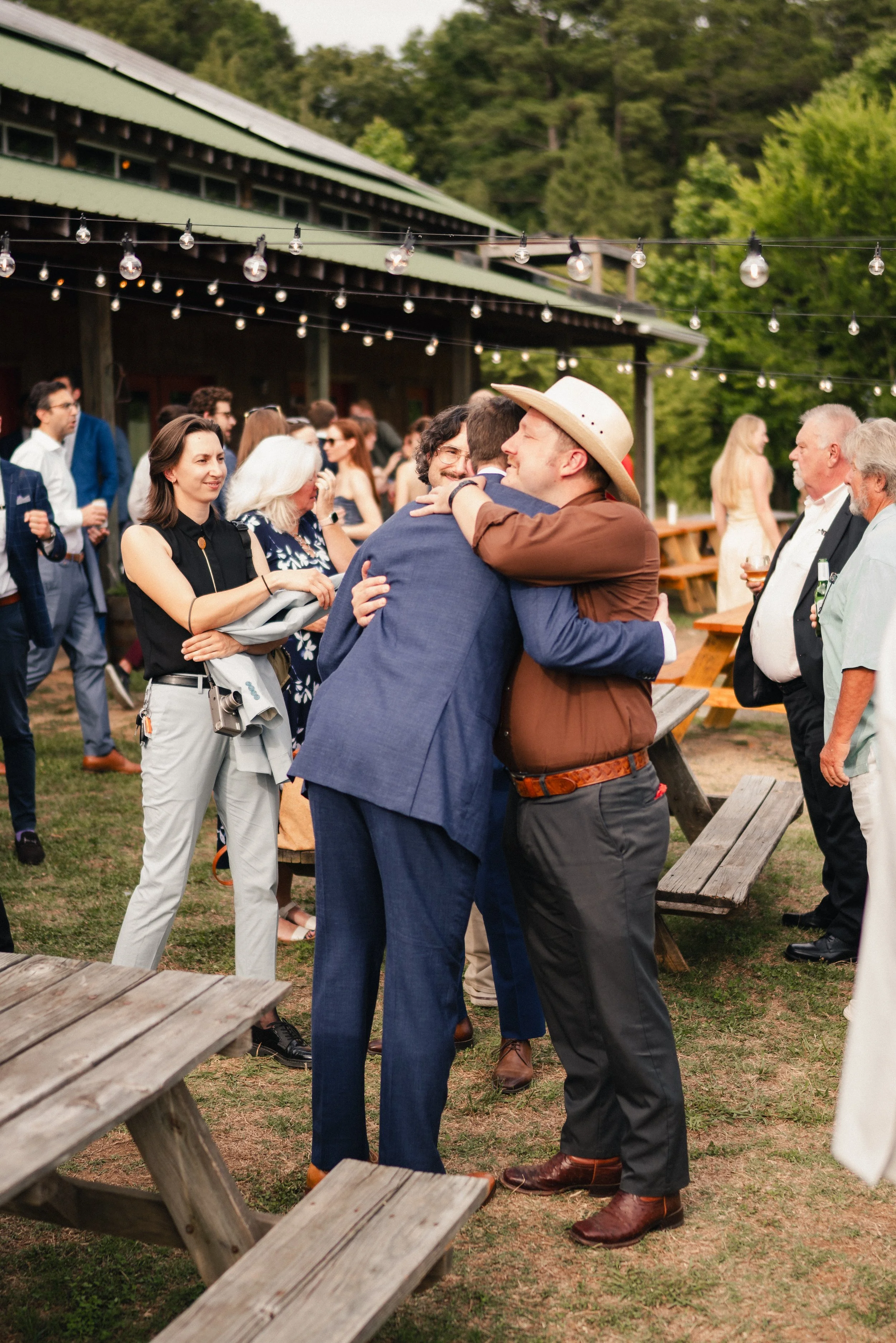 People gathering outdoors, hugging and socializing at a party under string lights with a green building and trees in the background.