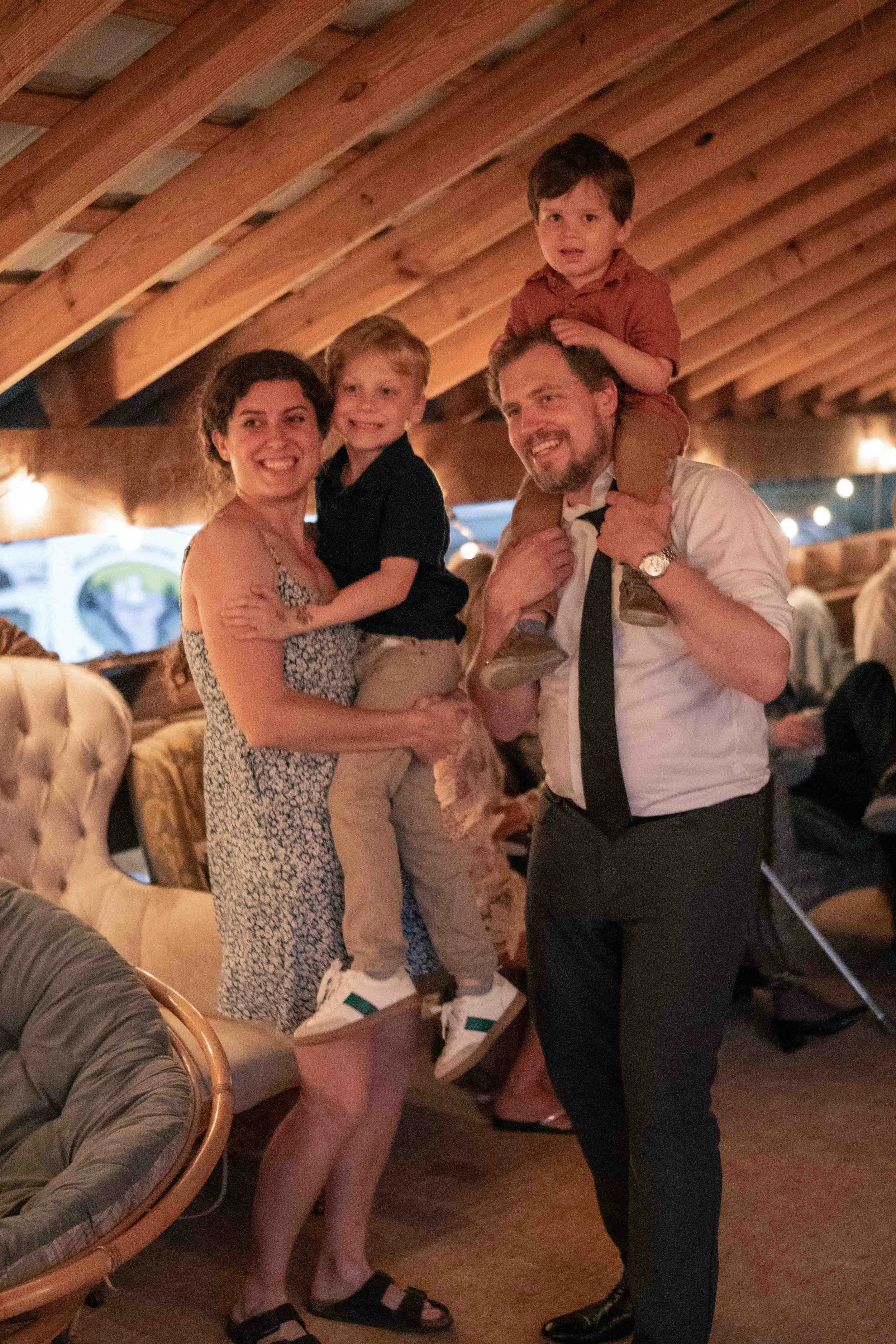 A family group of four people, two adults and two children, in a warmly lit restaurant or bar with wooden ceiling beams. The woman is holding a young boy, and the man is carrying a slightly older boy on his shoulders. They are smiling and enjoying th