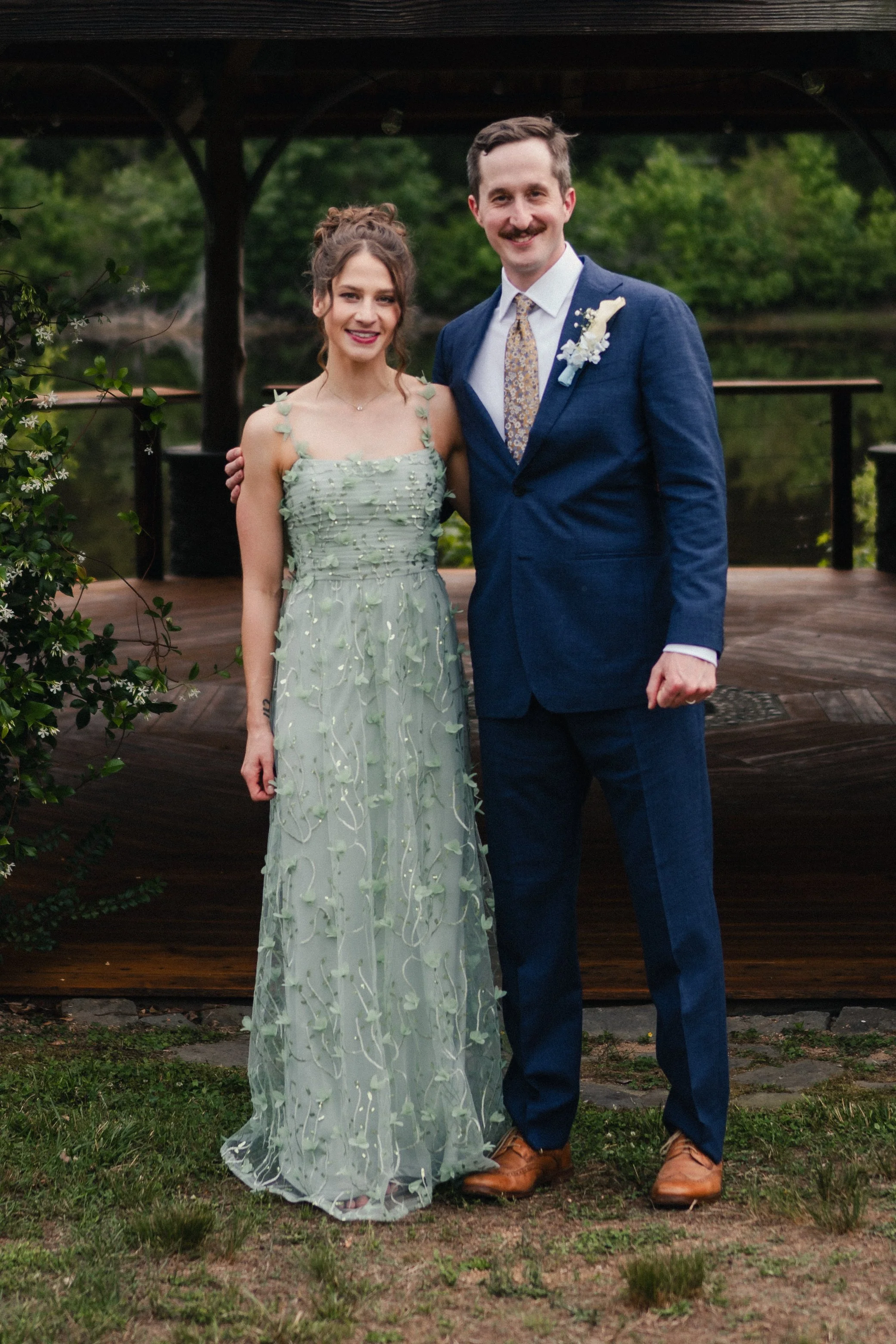 A man and woman pose together outdoors near a lake, with trees in the background. The woman wears a light green, floral, sleeveless dress, and the man wears a navy suit with a patterned tie. They are smiling and standing on grass.