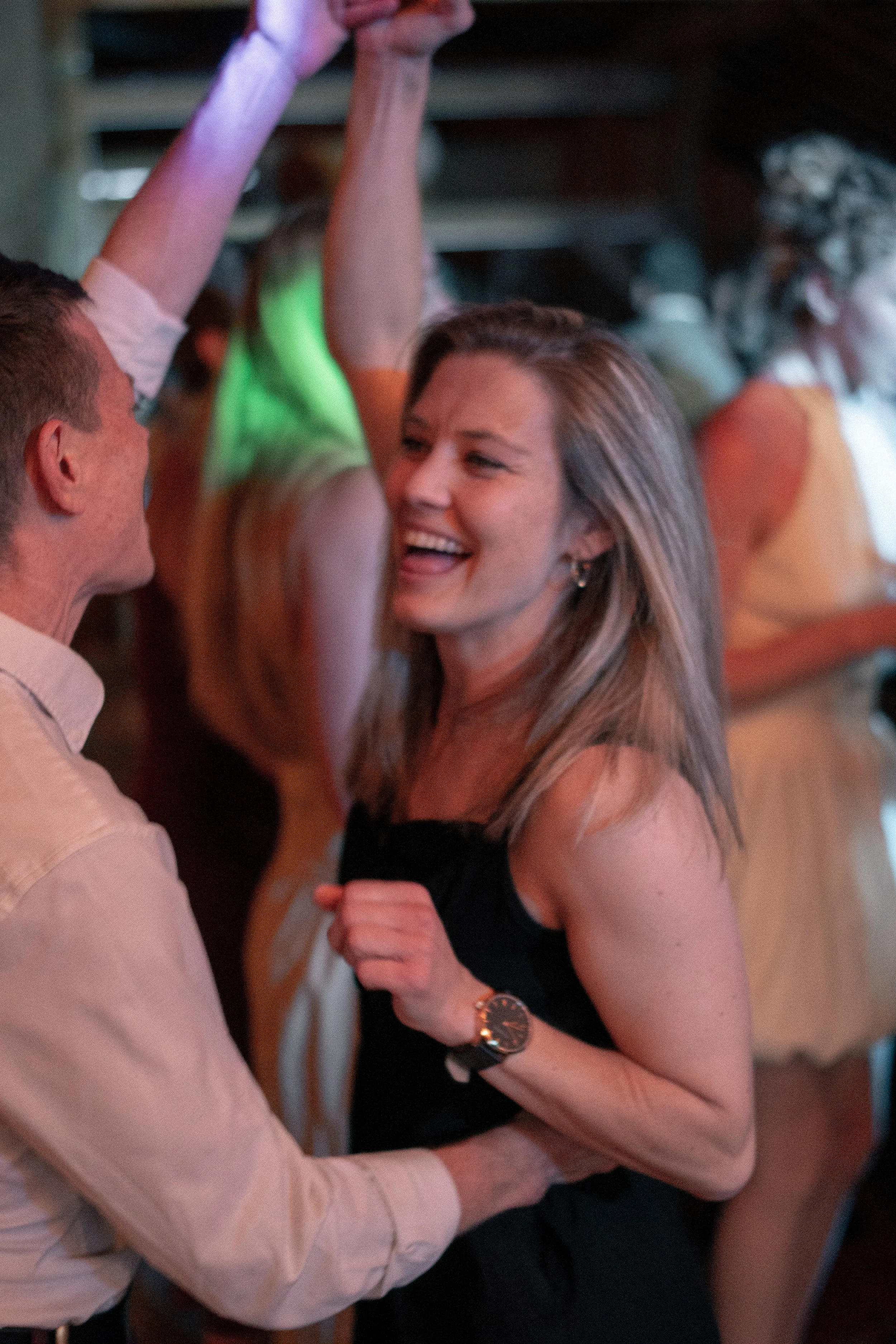A woman laughing and smiling while engaging in a dance with a man in a social setting. The woman has long hair, is wearing a black sleeveless top, and a wristwatch. The man is wearing a light-colored, long-sleeved shirt. Other people are visible in t
