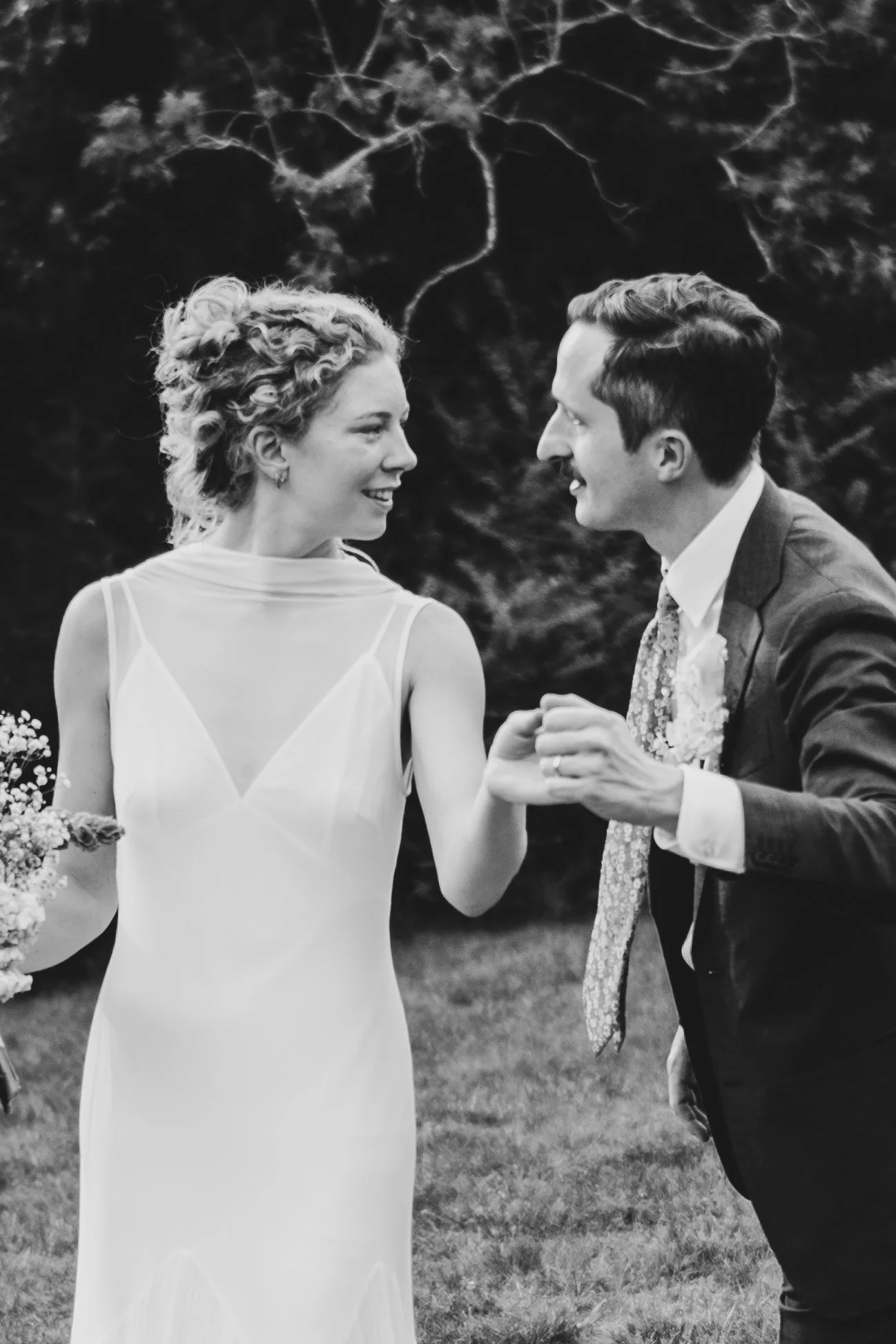 A bride and groom holding hands and smiling at each other during their wedding outdoors, with trees in the background, in black and white.