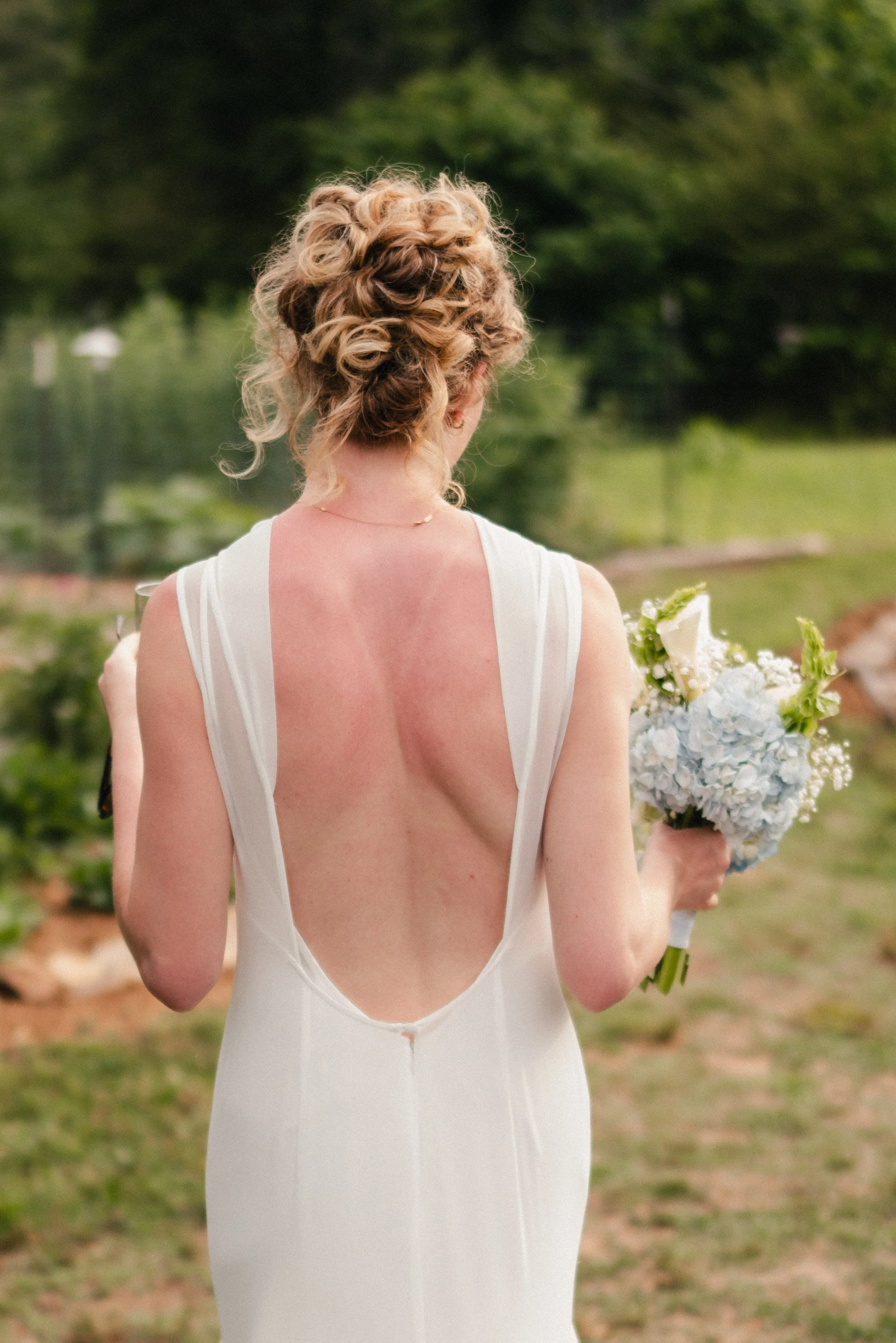 Back view of a woman in a white wedding dress holding a bouquet of white and green flowers, standing outdoors with a blurred green landscape in the background.