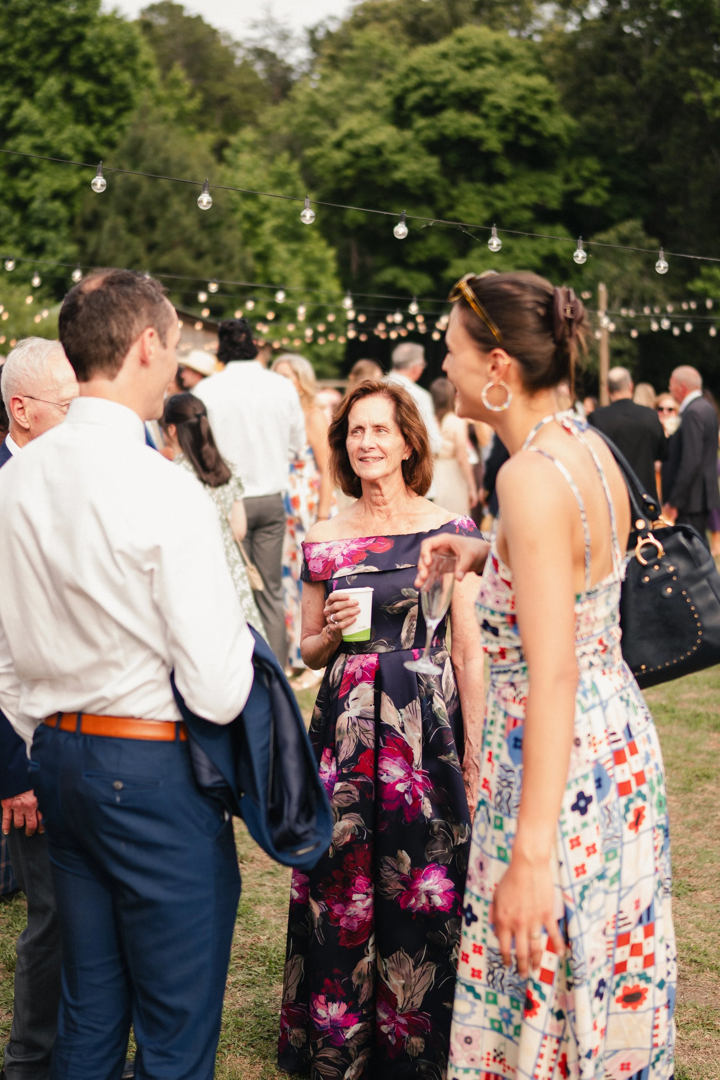 A group of people socializing outdoors during an event, with string lights hanging above and green trees in the background.