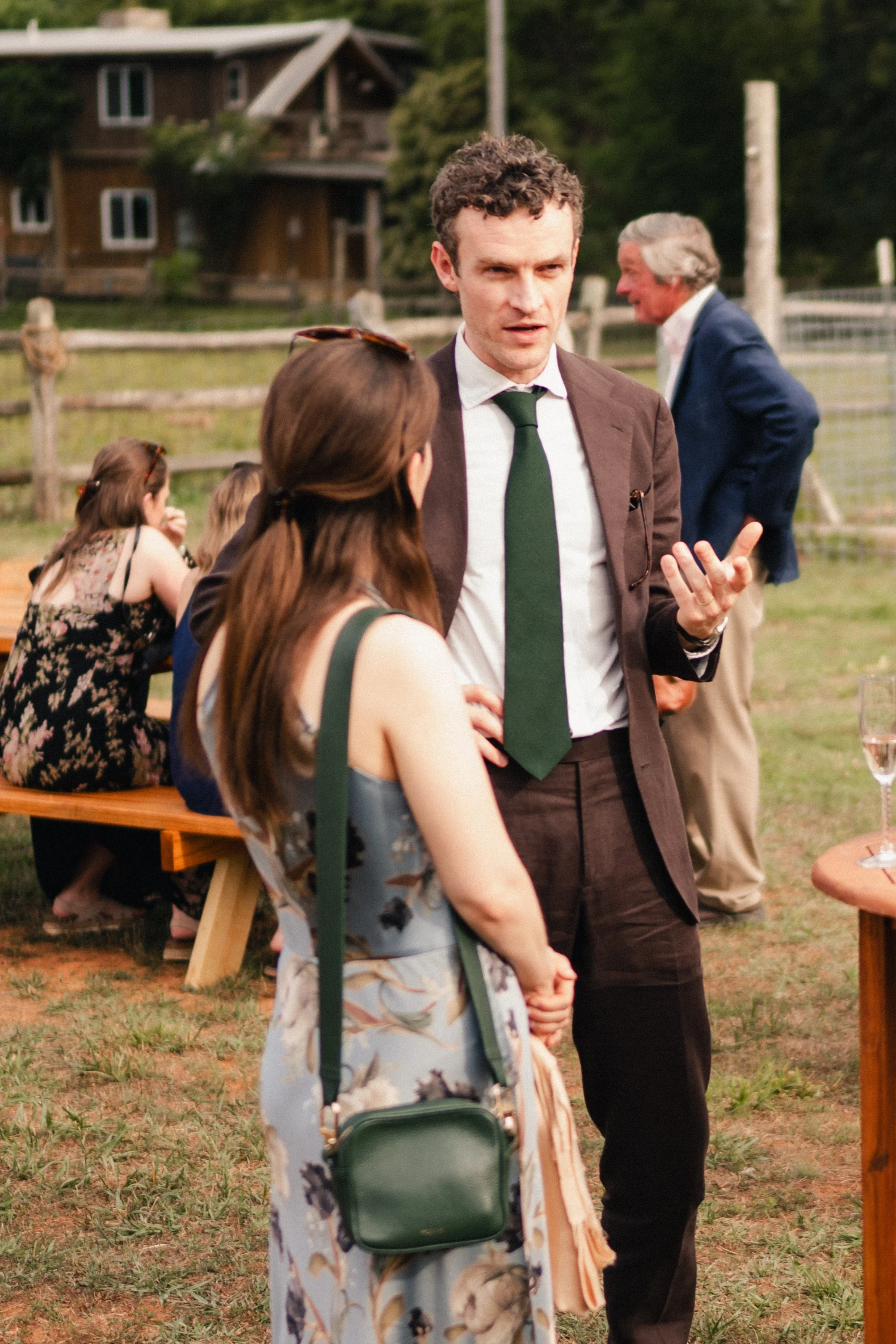 A man with curly hair wearing a brown suit and green tie is talking to a woman with long brown hair in a floral dress at an outdoor social event. In the background, another man in a navy suit is talking, and there are people sitting at a wooden picni