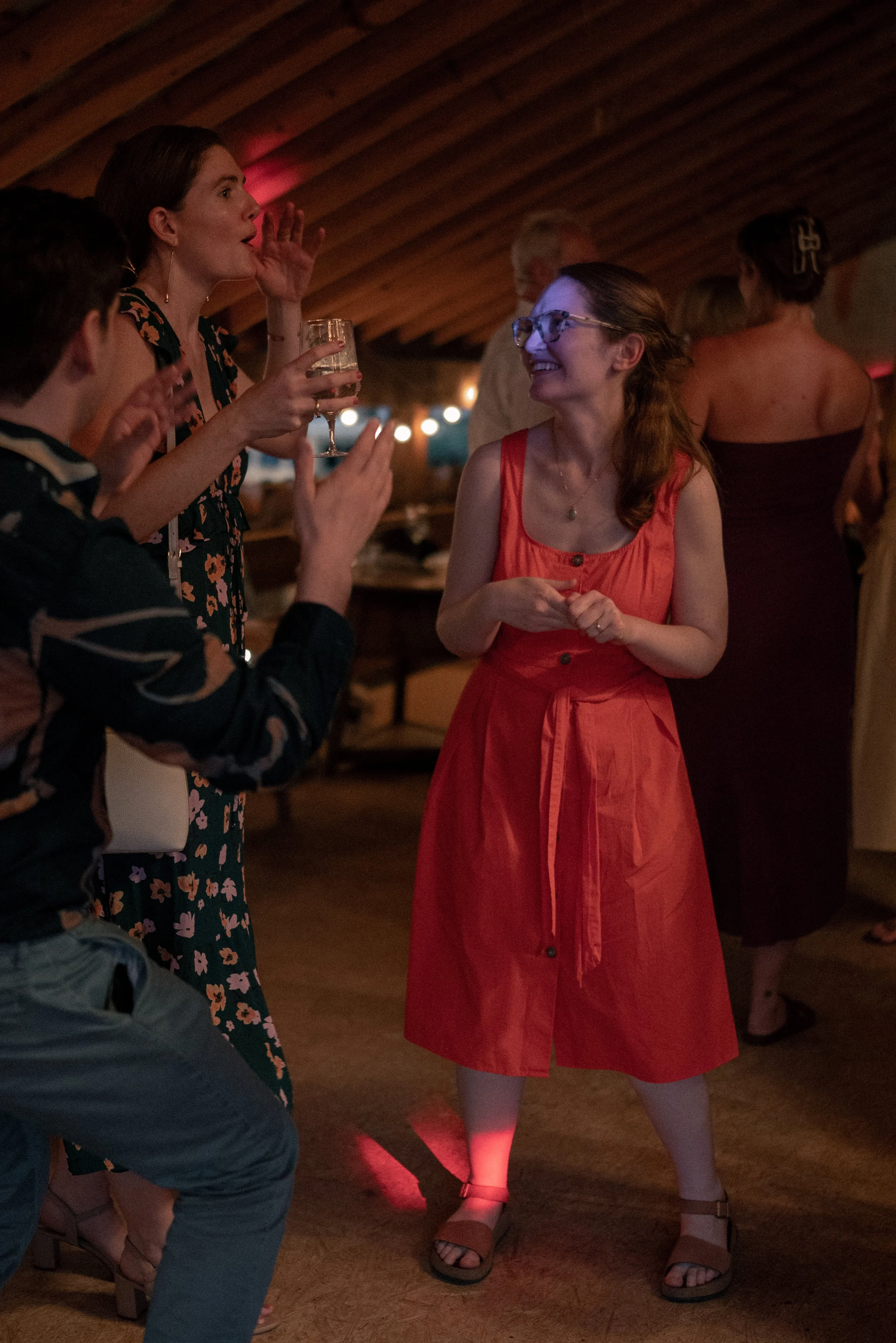 People socializing at a dimly lit indoor gathering, woman in red dress smiling at woman holding a glass of wine, others in casual attire.