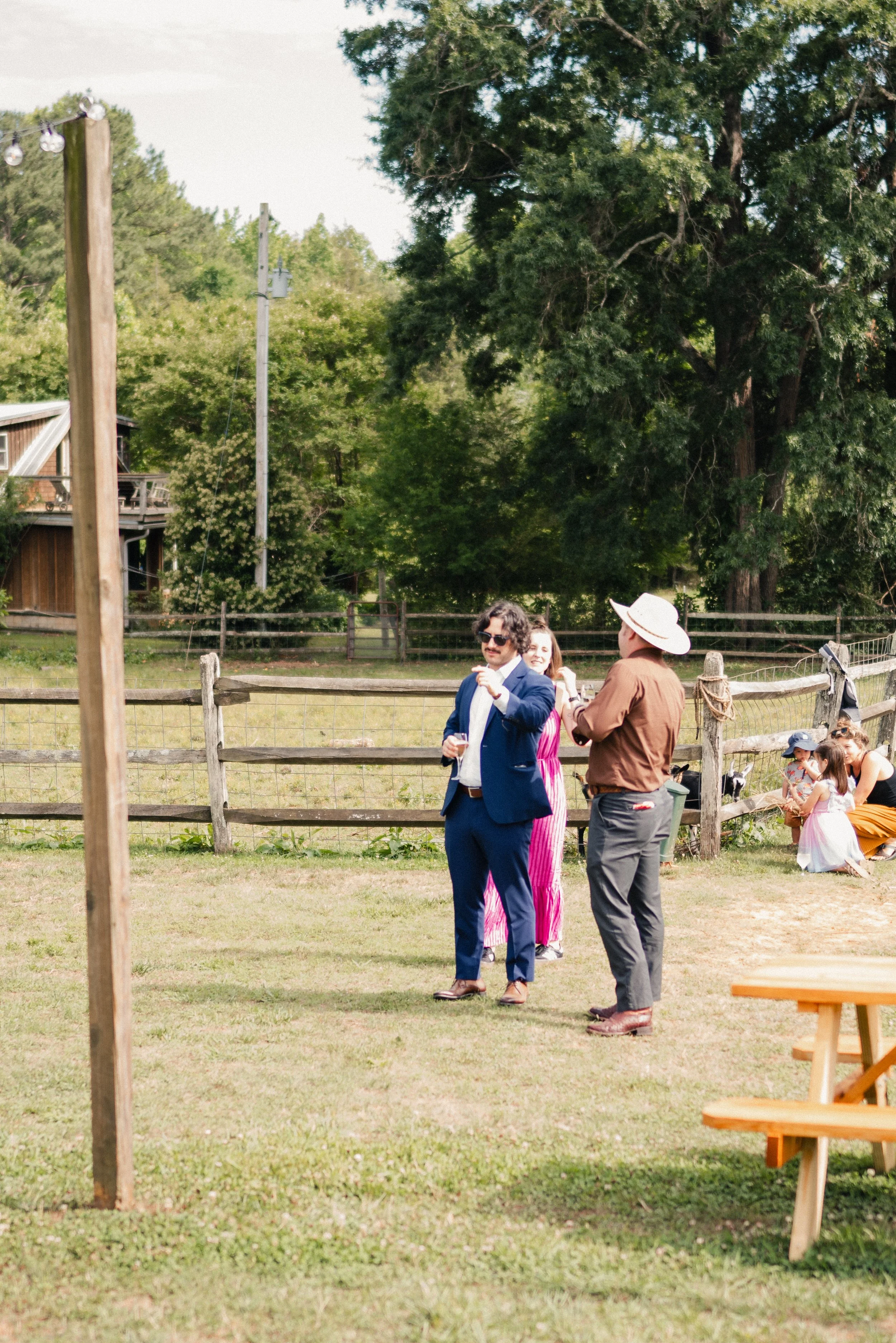 People dressed in formal and casual attire standing and talking outdoors in a grassy area with trees and a wooden fence, possibly at a social event or gathering.