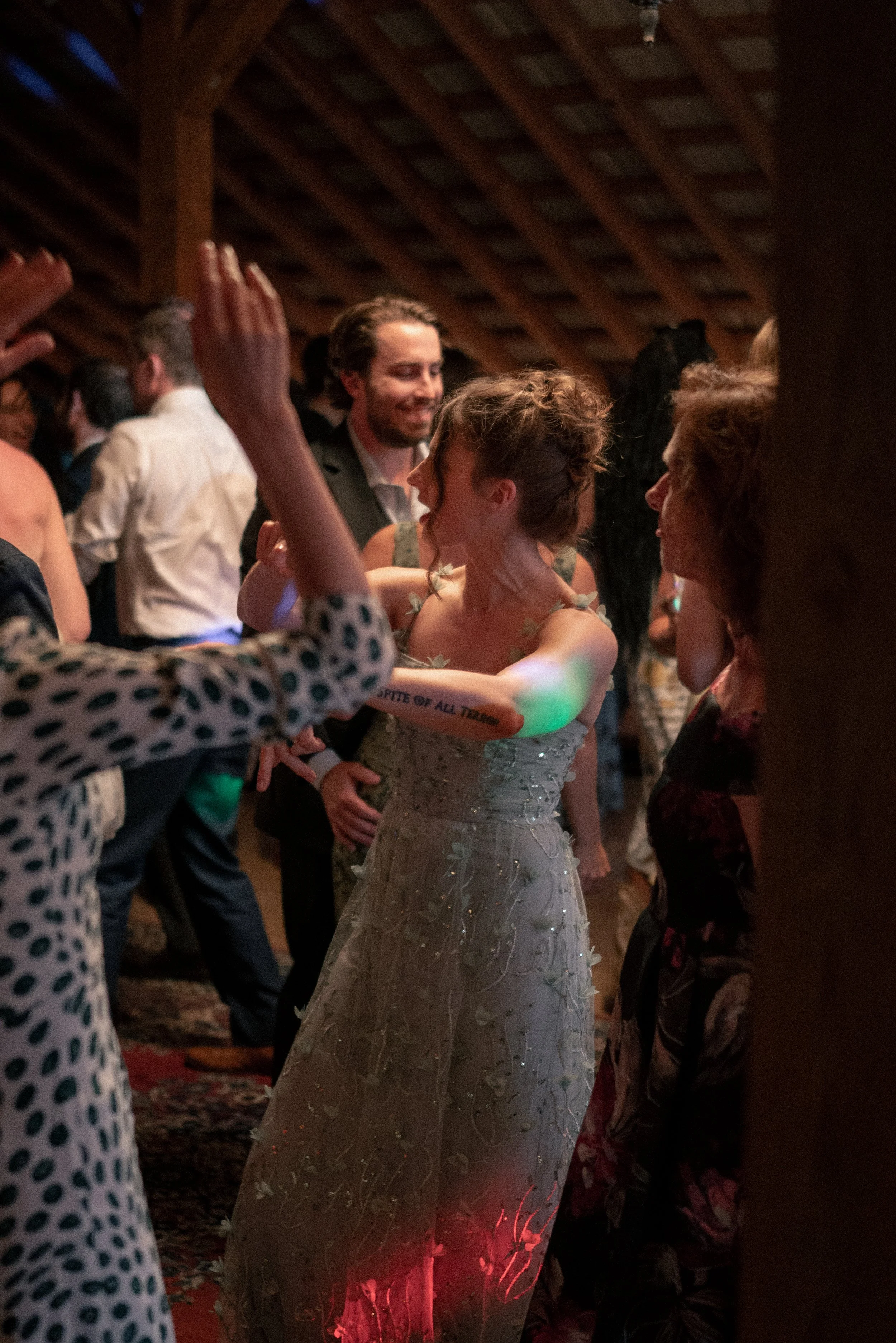 People dancing closely at a social event in a dimly lit room with wooden beams on the ceiling. The focus is on a woman in a white dress with floral details and a tattoo on her arm, dancing with a man smiling in the background. Other guests are visibl