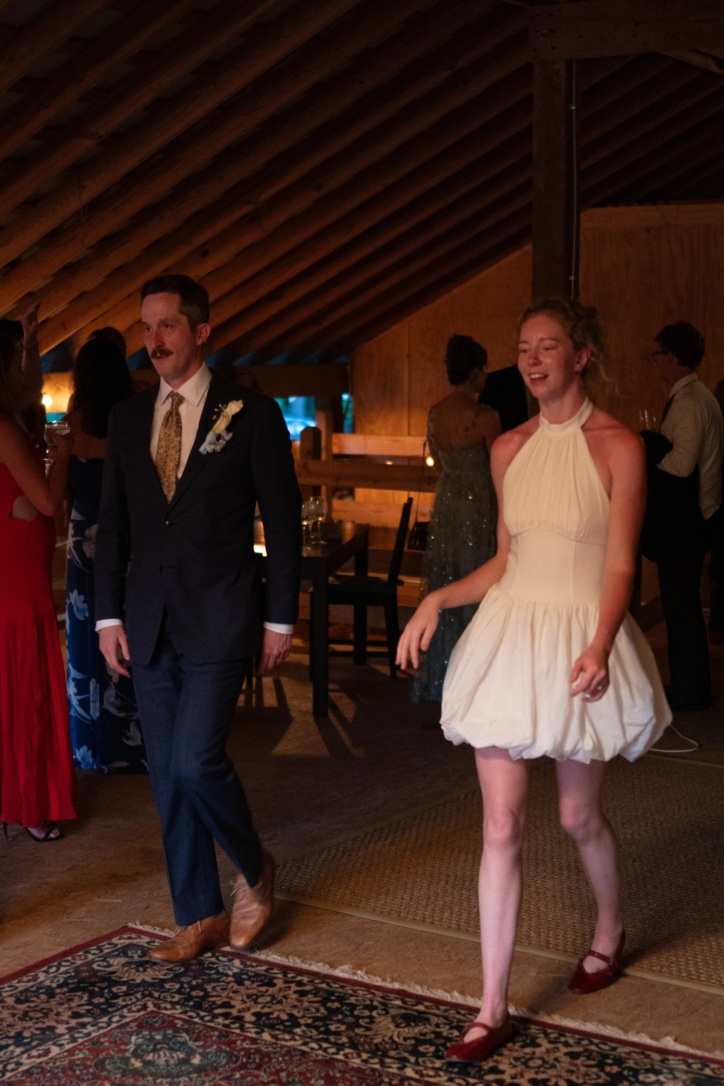 A man in a dark suit with a boutonniere and a woman in a cream-colored dress are at a celebration or party. Other people are in the background, some dancing or talking, in a warmly lit room with a wooden ceiling and carpeted floor.
