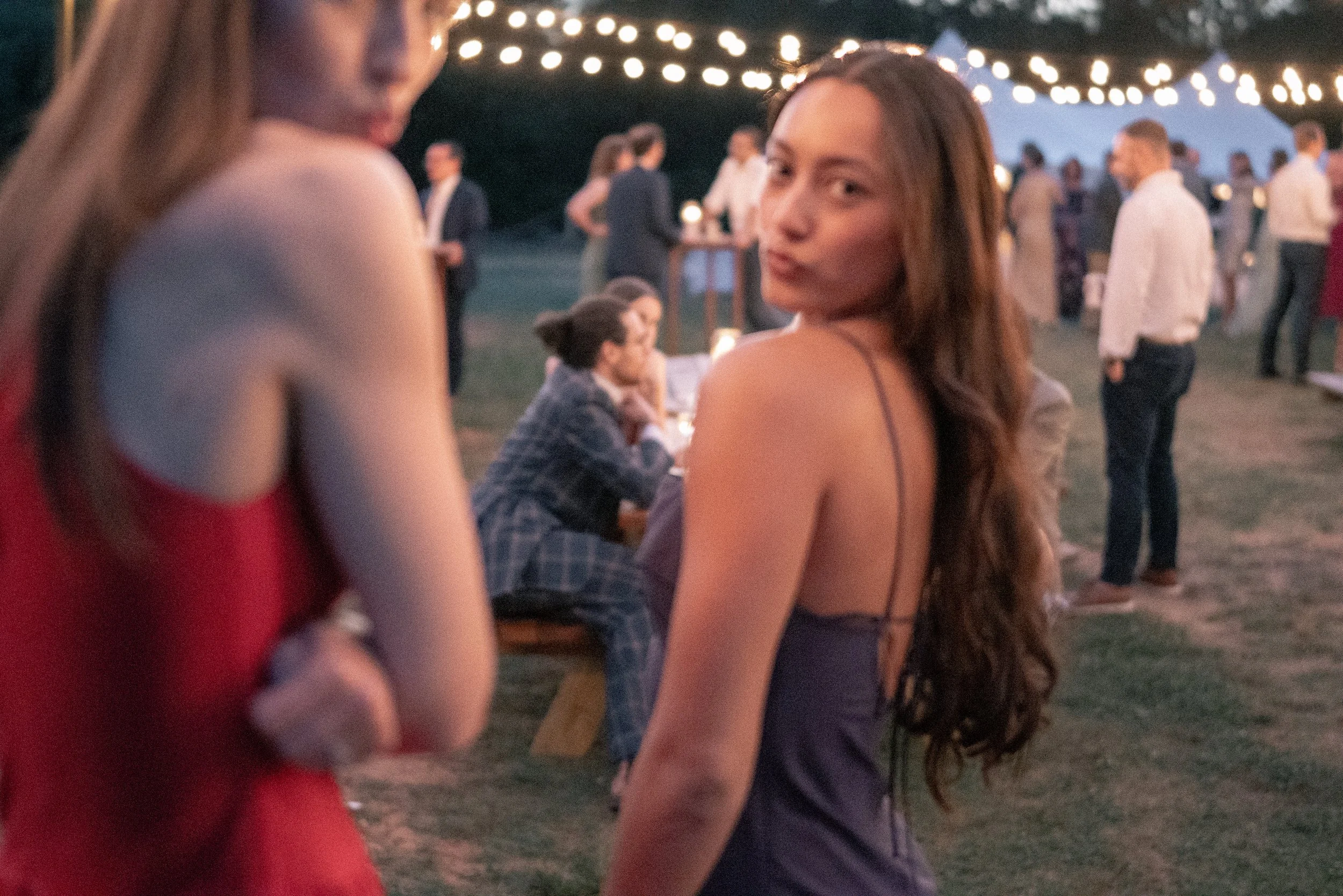 Two women dancing at an outdoor evening event with string lights overhead, with blurred guests in the background.