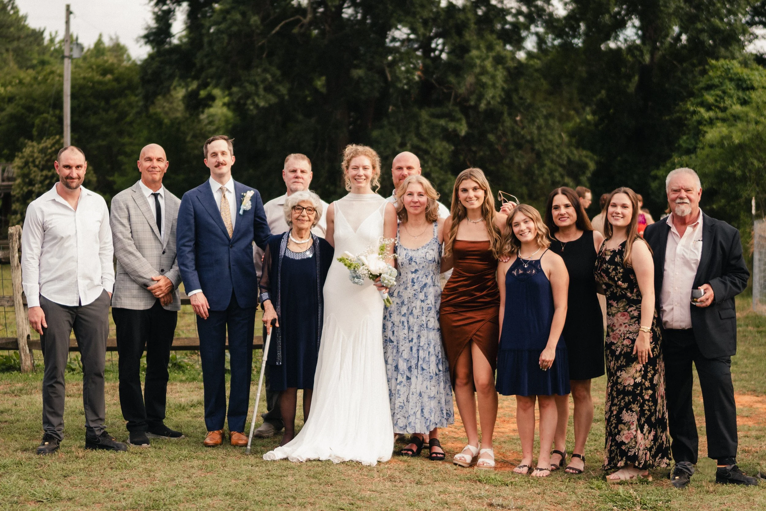 Group of 13 people, including a bride in a white dress holding a bouquet, and a groom in a blue suit, posing outdoors on a grassy area with trees in the background, at a wedding celebration.