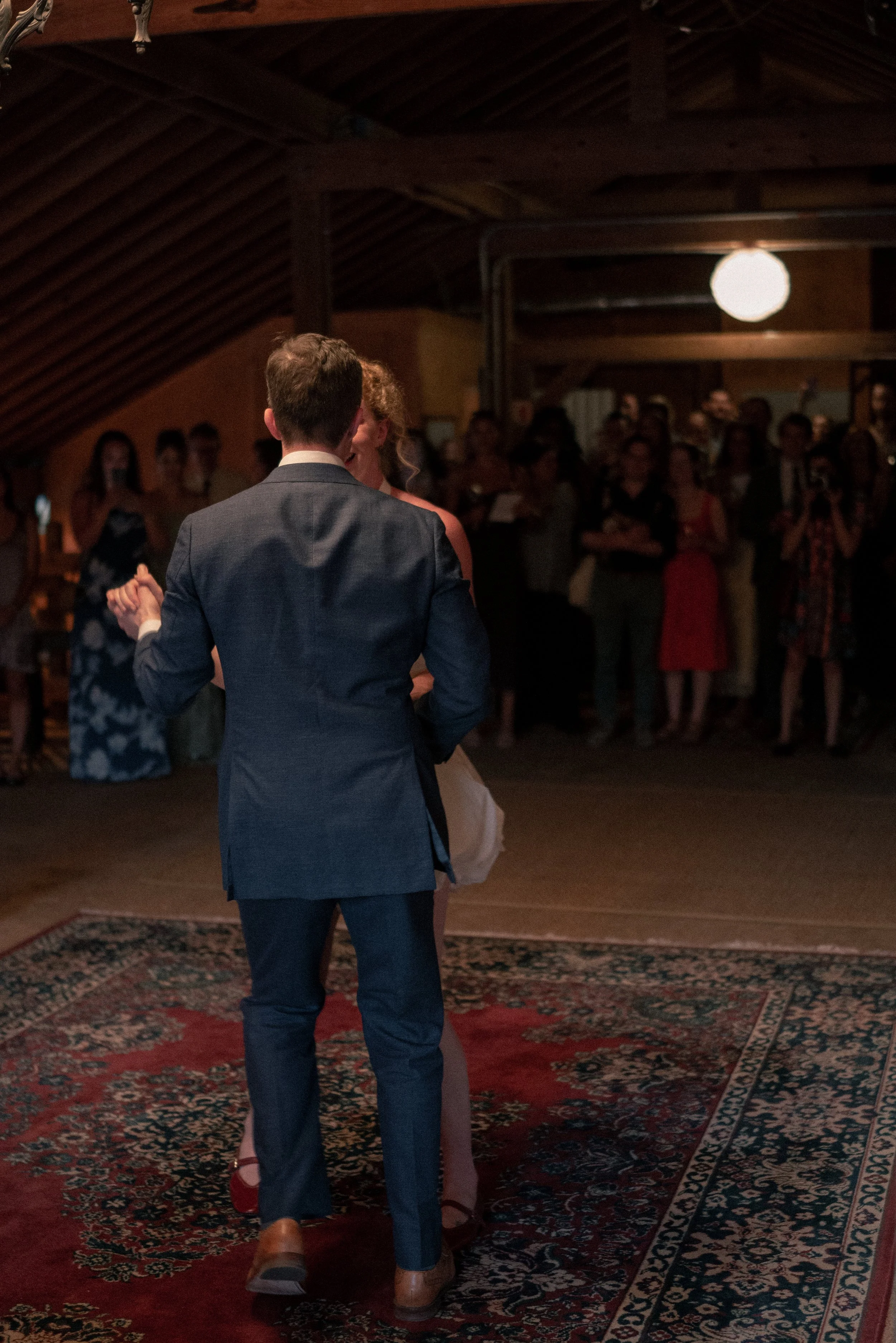 A bride and groom dancing at their wedding reception with guests watching in the background. The scene is set in a rustic venue with wooden ceiling beams and a patterned rug on the floor.