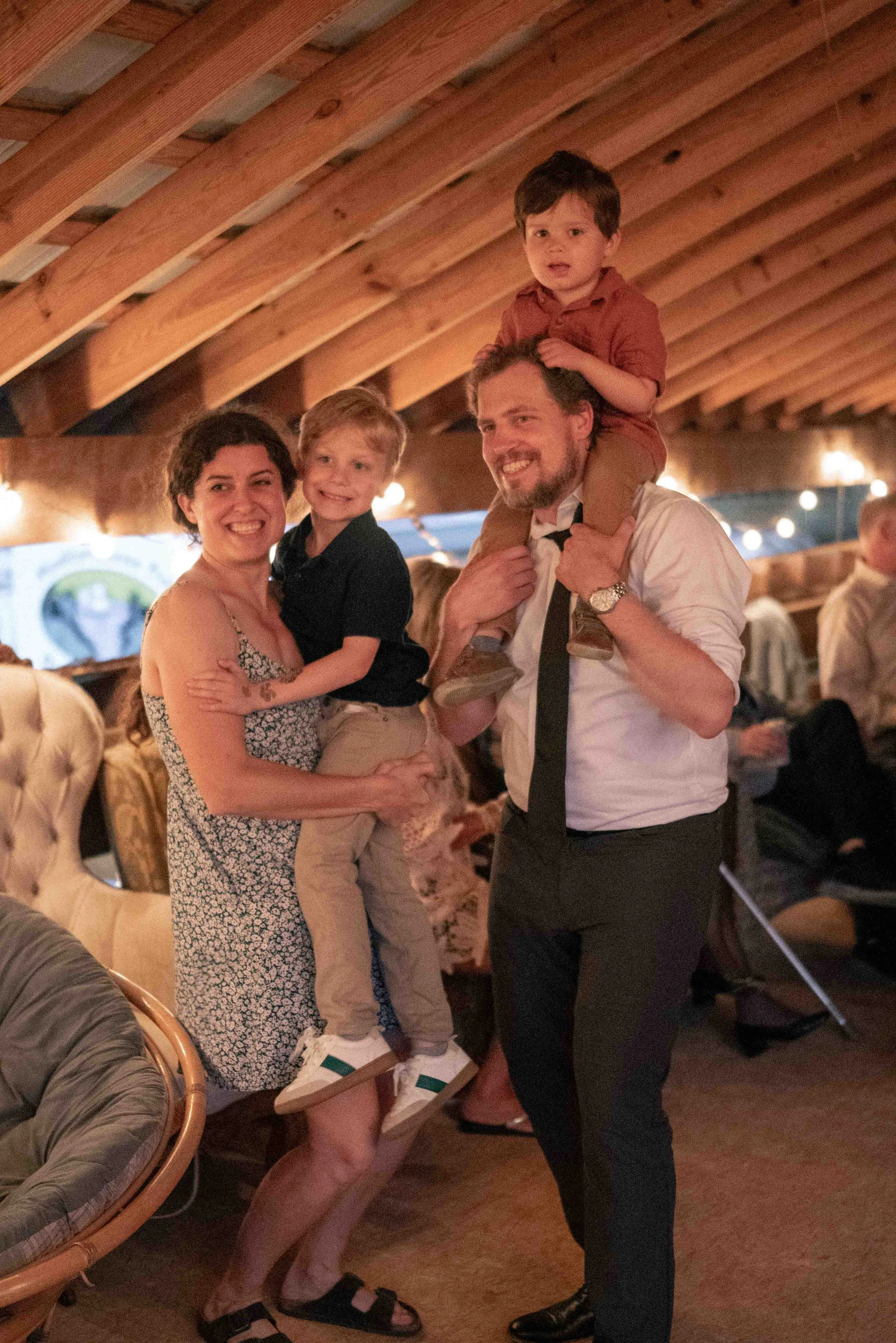 A happy family of four, including a woman, a man, and two young boys, gathered together at a restaurant or event space with wood-paneled ceiling. The man is giving the younger boy a piggyback ride, and the woman is holding the older boy. Everyone is 