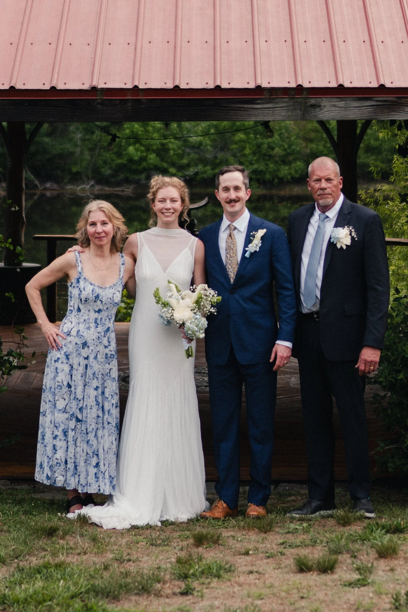 A group of four people dressed in formal attire, standing outdoors in front of a body of water and greenery, likely at a wedding.