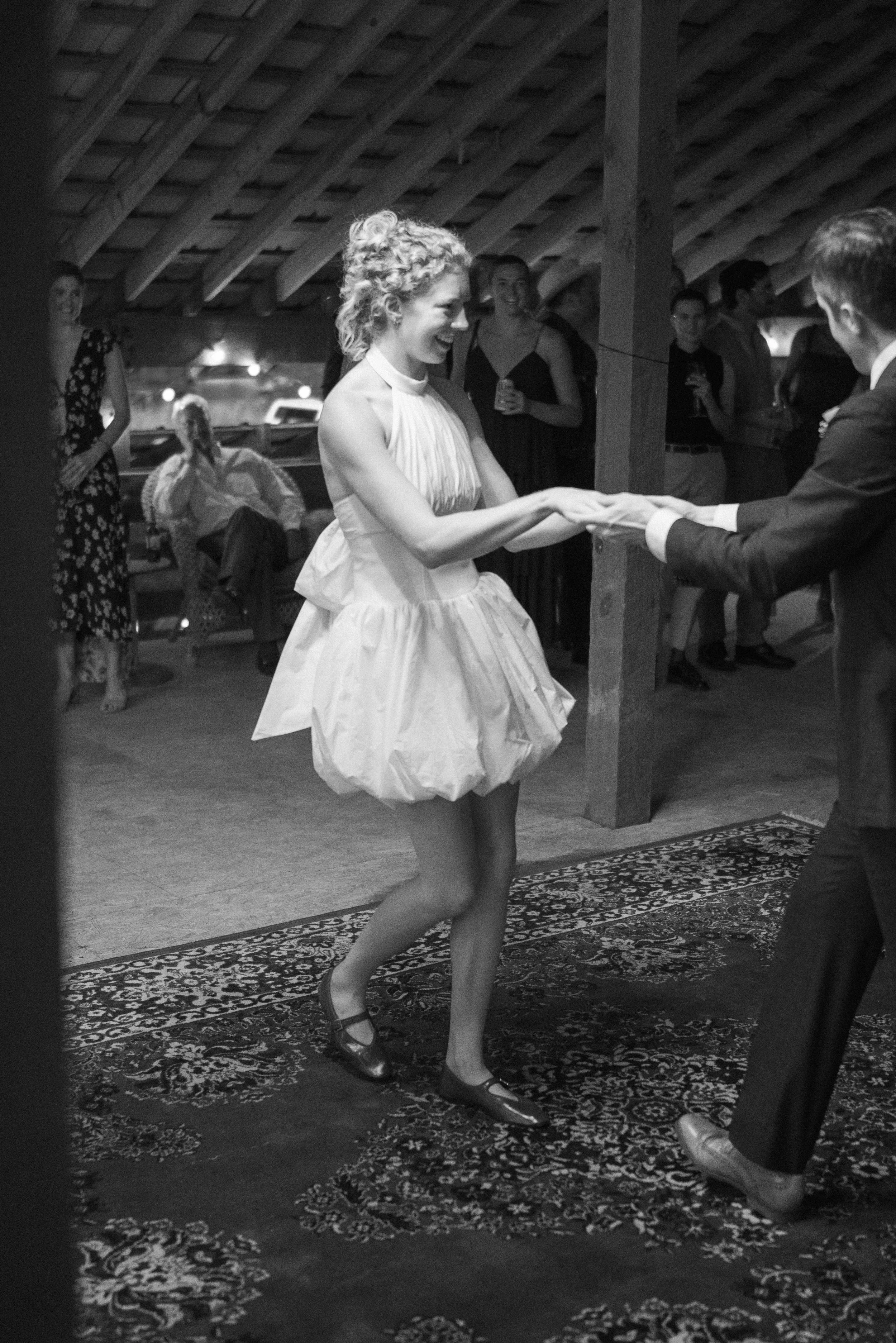 A woman in a white dress and ballet shoes dancing with a man in a suit, surrounded by onlookers at an indoor event in a rustic venue.
