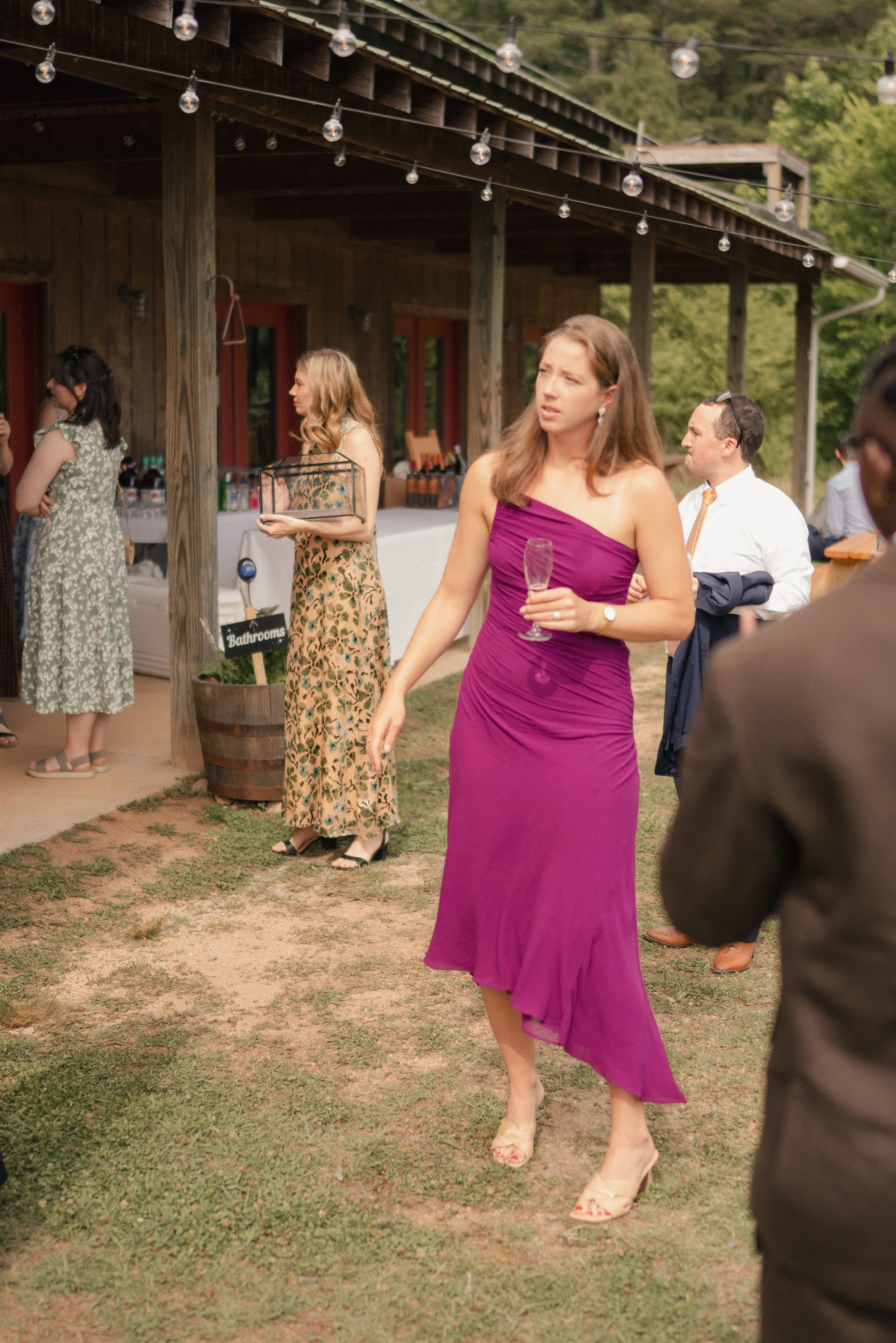 Women dressed in formal attire at an outdoor event, standing on a grassy area with string lights overhead and a wooden building in the background.