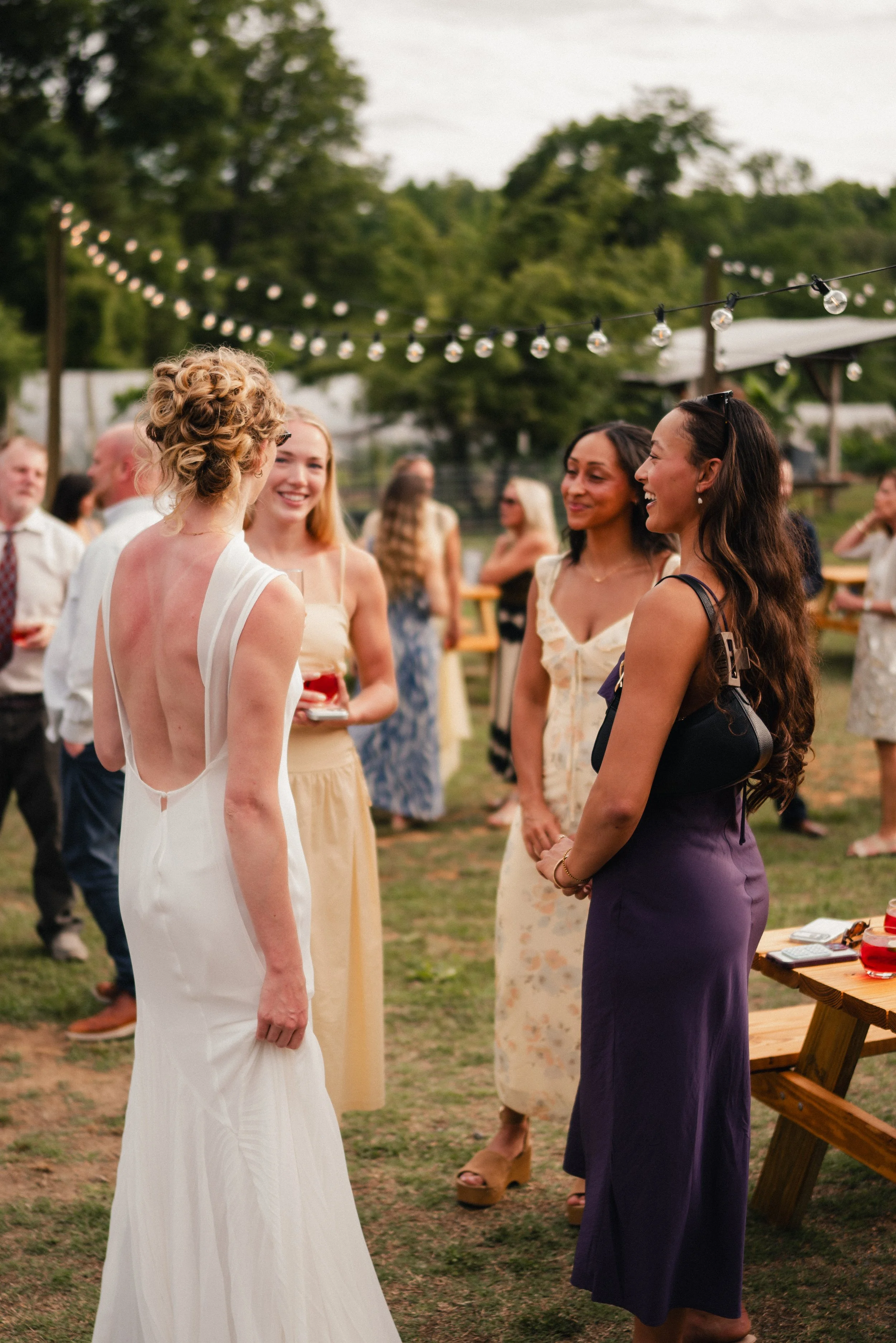 At an outdoor wedding reception, a bride dressed in white is facing women who are smiling and talking to her. The women are wearing colorful dresses, and the scene is decorated with string lights hanging overhead. There are trees and other guests in 