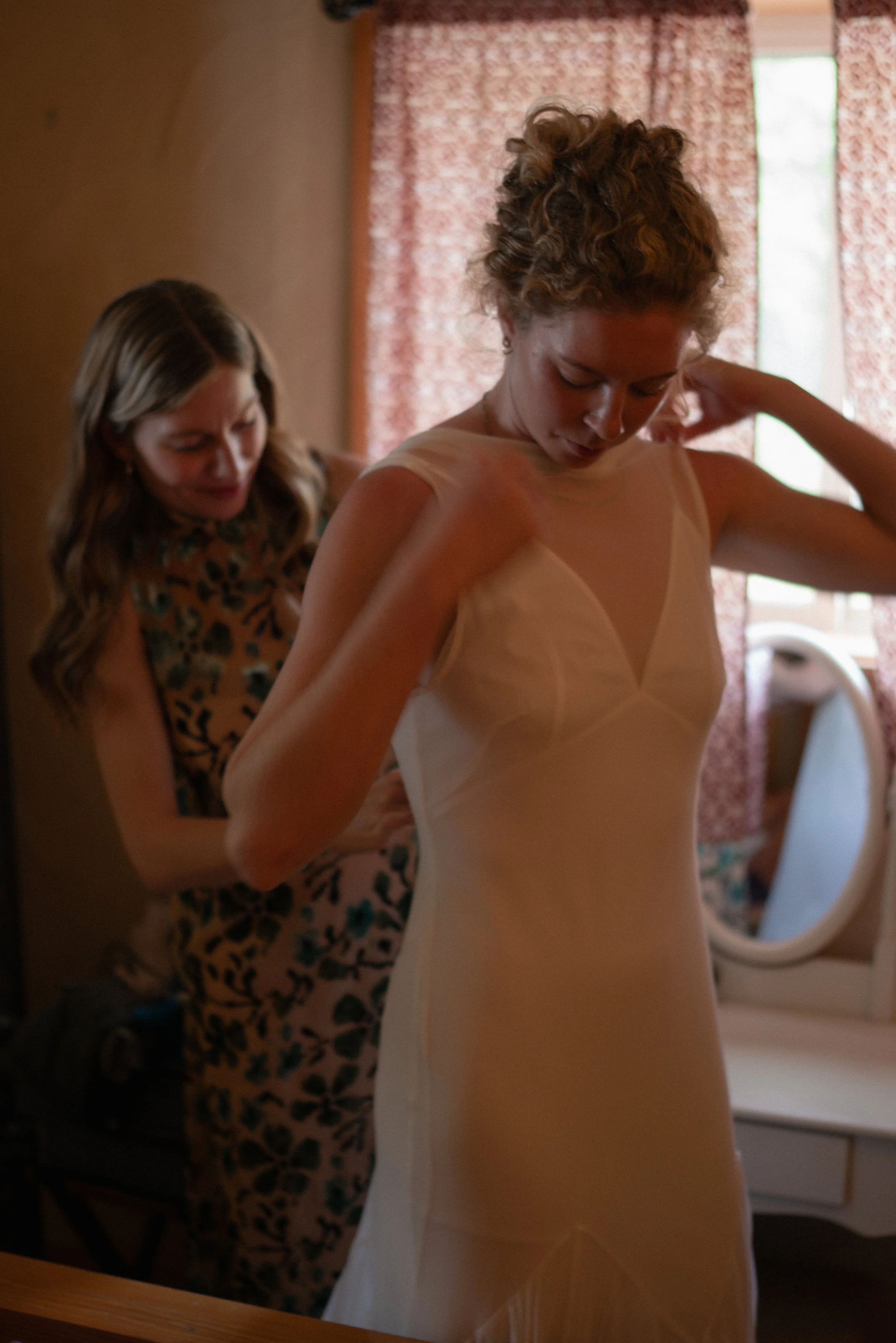 A woman with curly hair standing in front of a mirror, adjusting her dress, with another woman helping her in a softly lit room with pink curtains in the background.