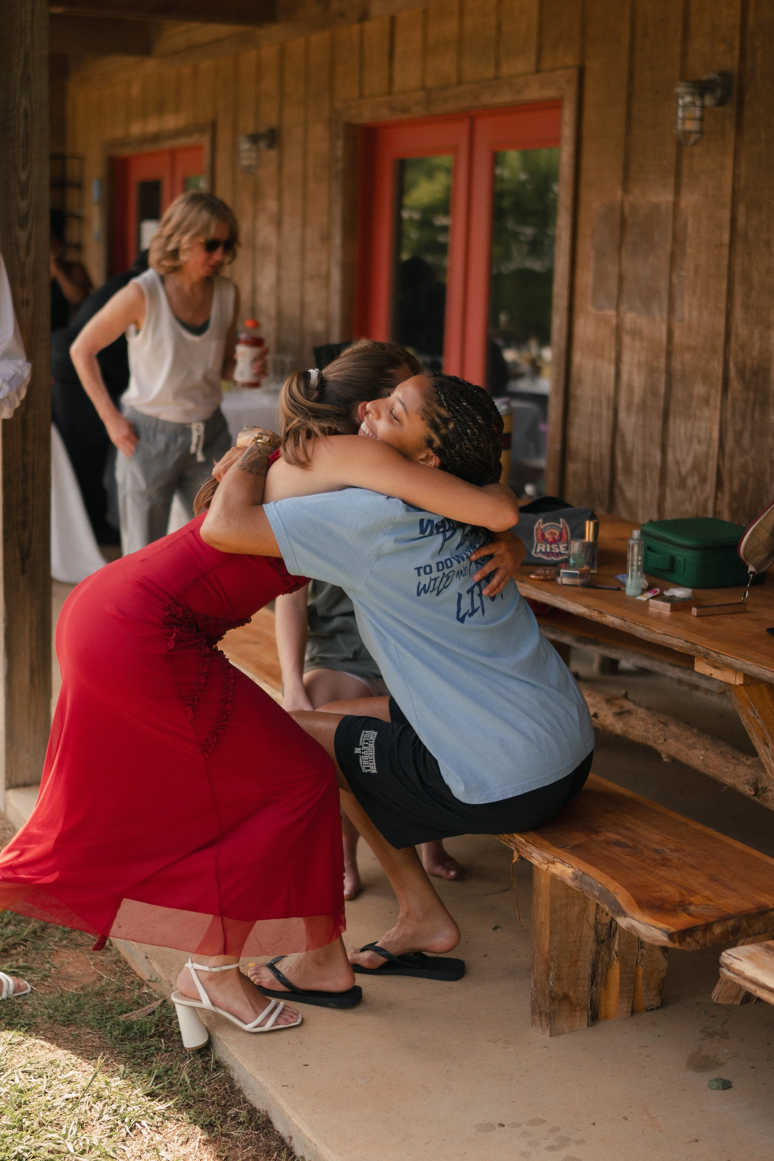 Two women embrace in a hug near a wooden picnic table outside, one with long dark hair and the other with braids, smiling. A third woman in sunglasses and a white tank top stands nearby, and a fourth woman seated at the table is visible behind them. 