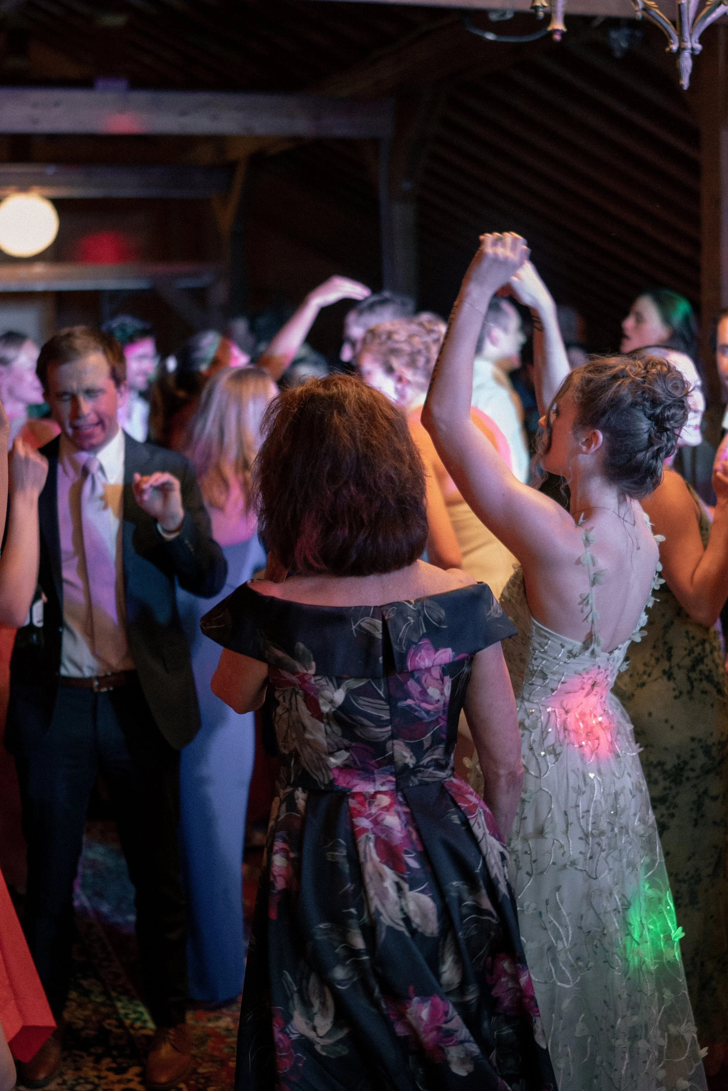 A group of people dancing at a party or celebration. The scene is lively with a mix of men and women in formal and semi-formal attire, including dresses and suits. The setting appears to be a decorated hall with dim lighting.