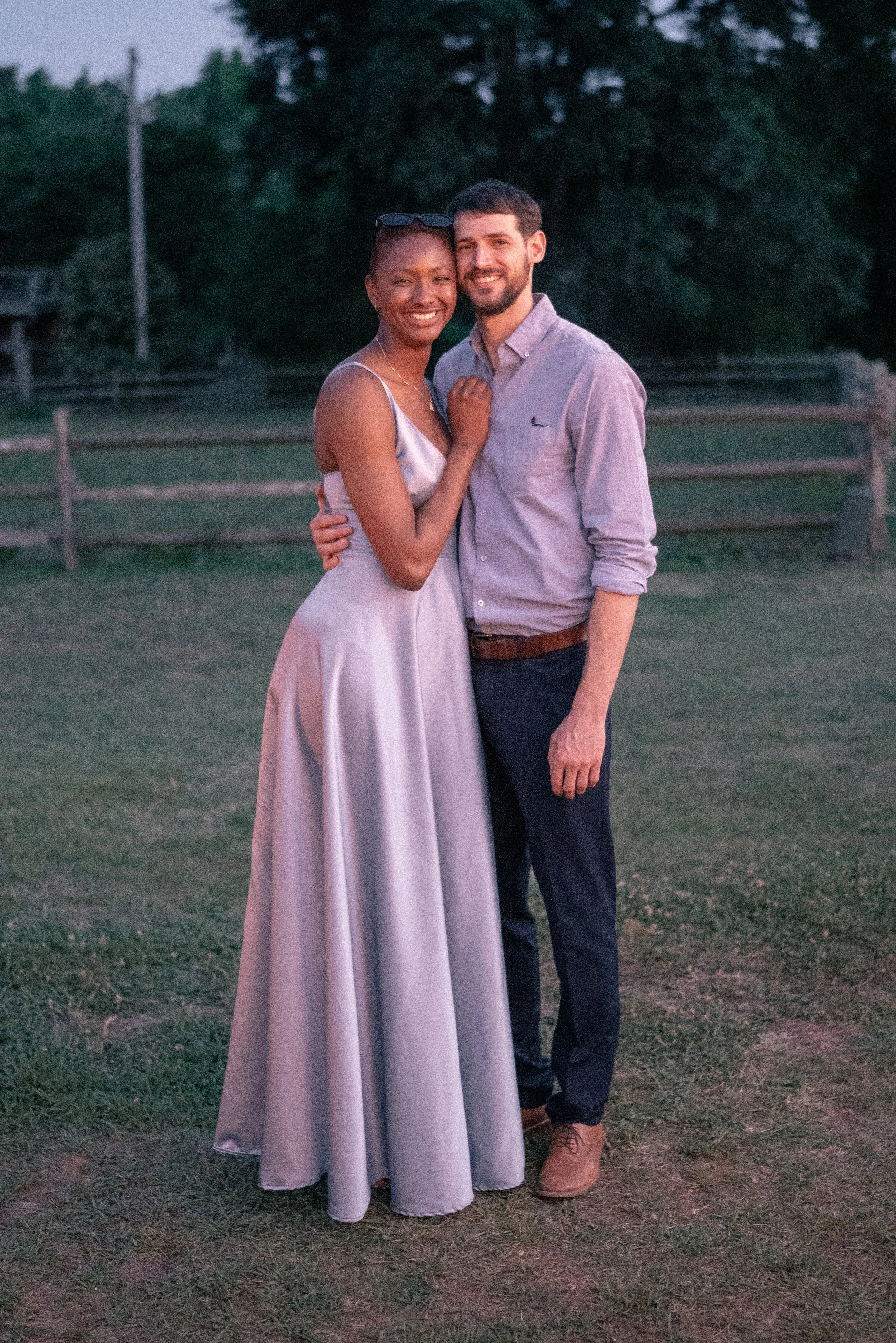 A happy couple standing outdoors on a grassy field during dusk. The woman is wearing a light purple satin dress and sunglasses on her head, while the man is dressed in a light pink button-up shirt and dark pants. They are smiling and embracing each o