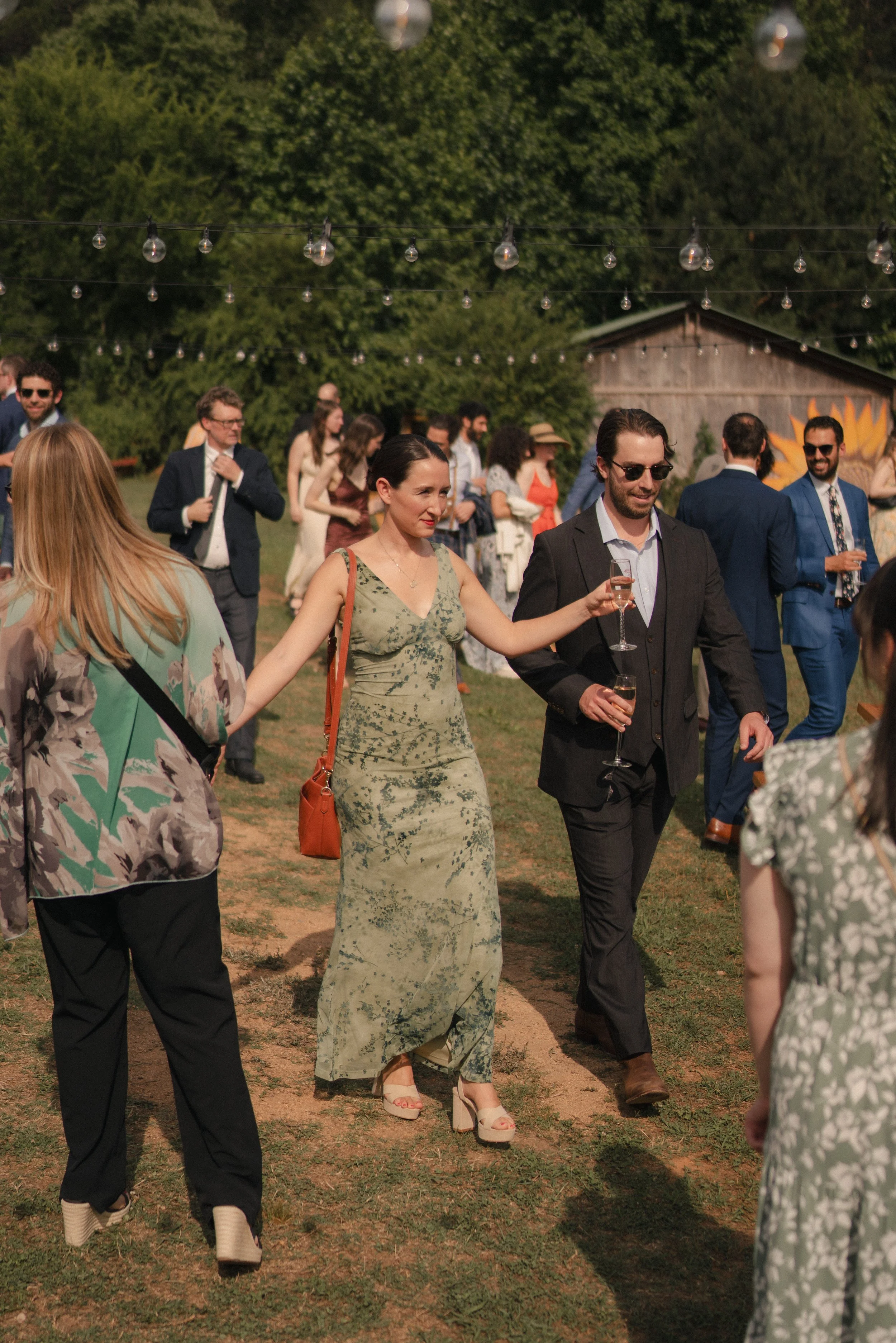 People dancing outdoors at a social gathering during the late afternoon or early evening, with string lights hanging overhead and a rustic building with a mural in the background.