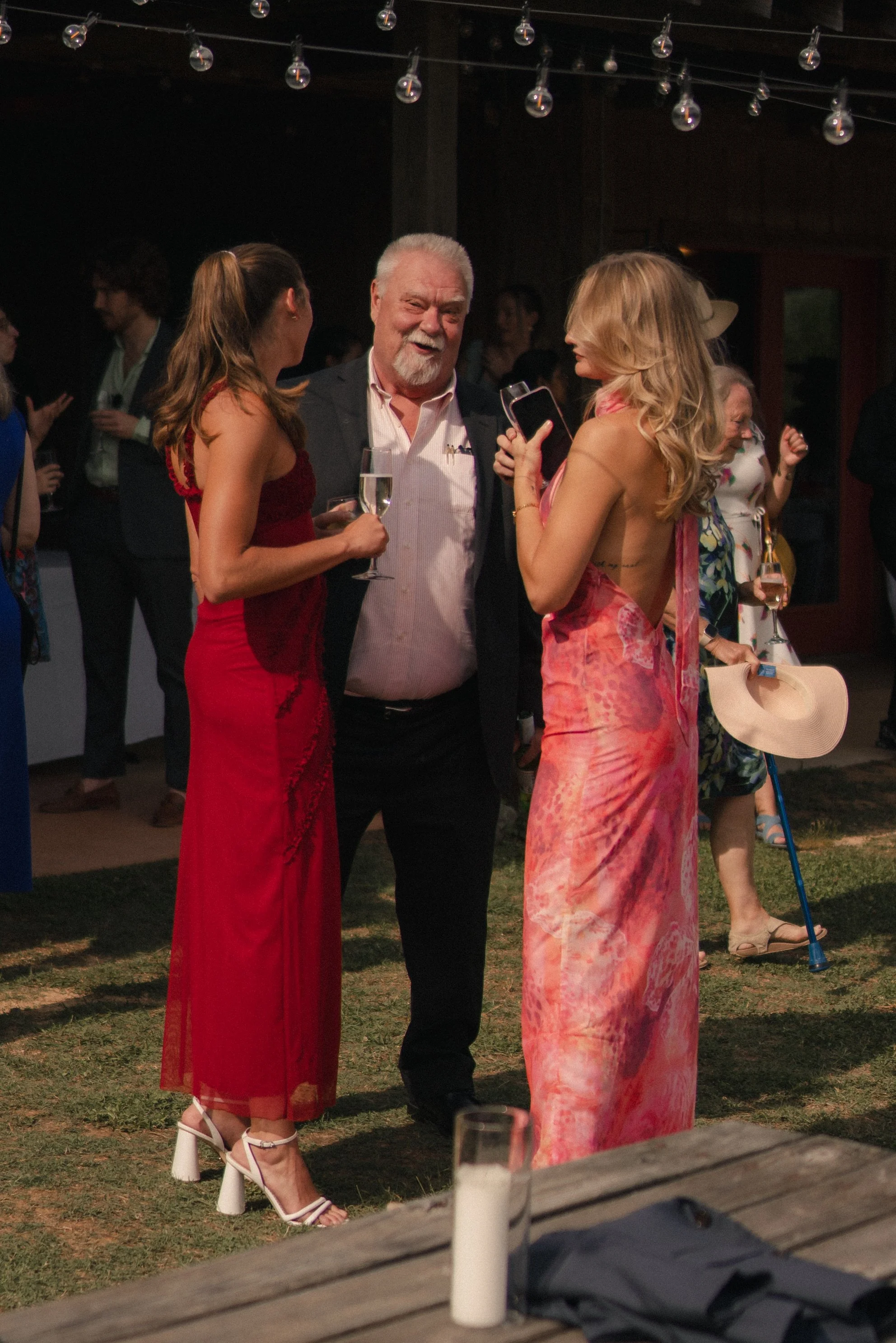 People at a social gathering, a woman in a red dress and white heels talking to a woman in a pink dress, a man in a dark jacket and white shirt holding a champagne glass, other guests in the background, and string lights above.