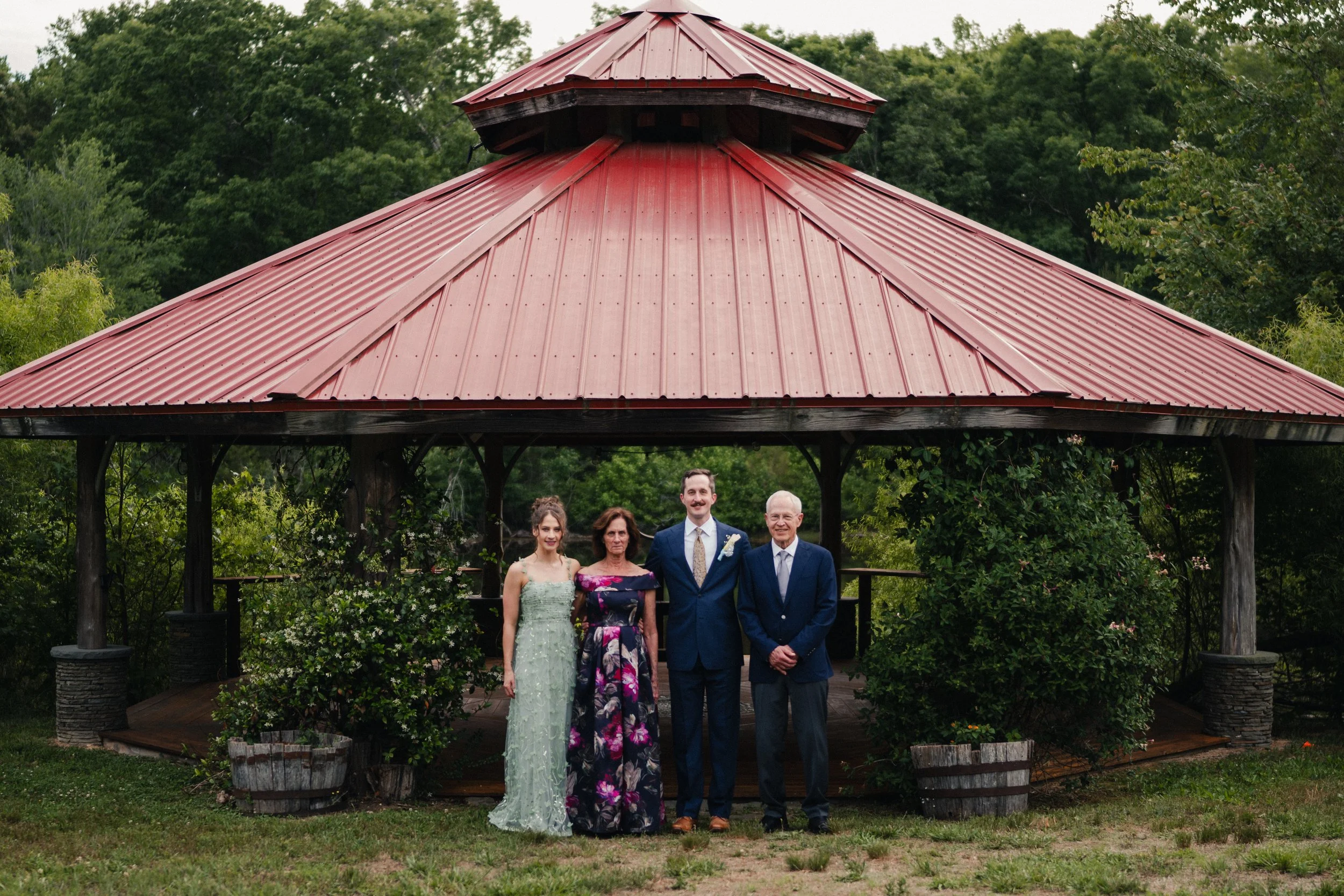 Group of five people standing under a gazebo with a red metal roof, surrounded by trees and greenery at an outdoor event or gathering.