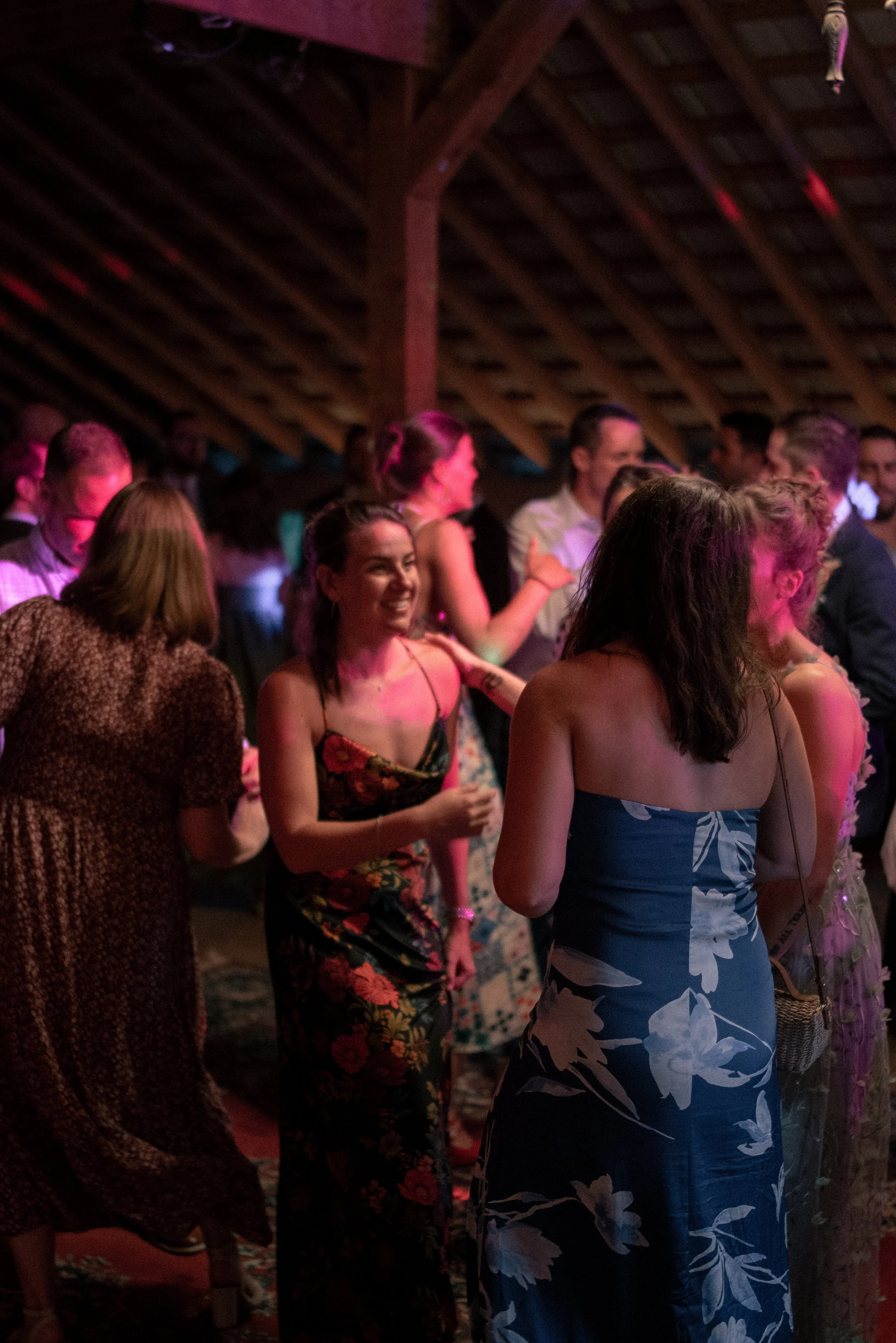 Group of people socializing at a party in a room with wooden ceiling beams. Some women are dressed in floral and strapless dresses, smiling and talking.