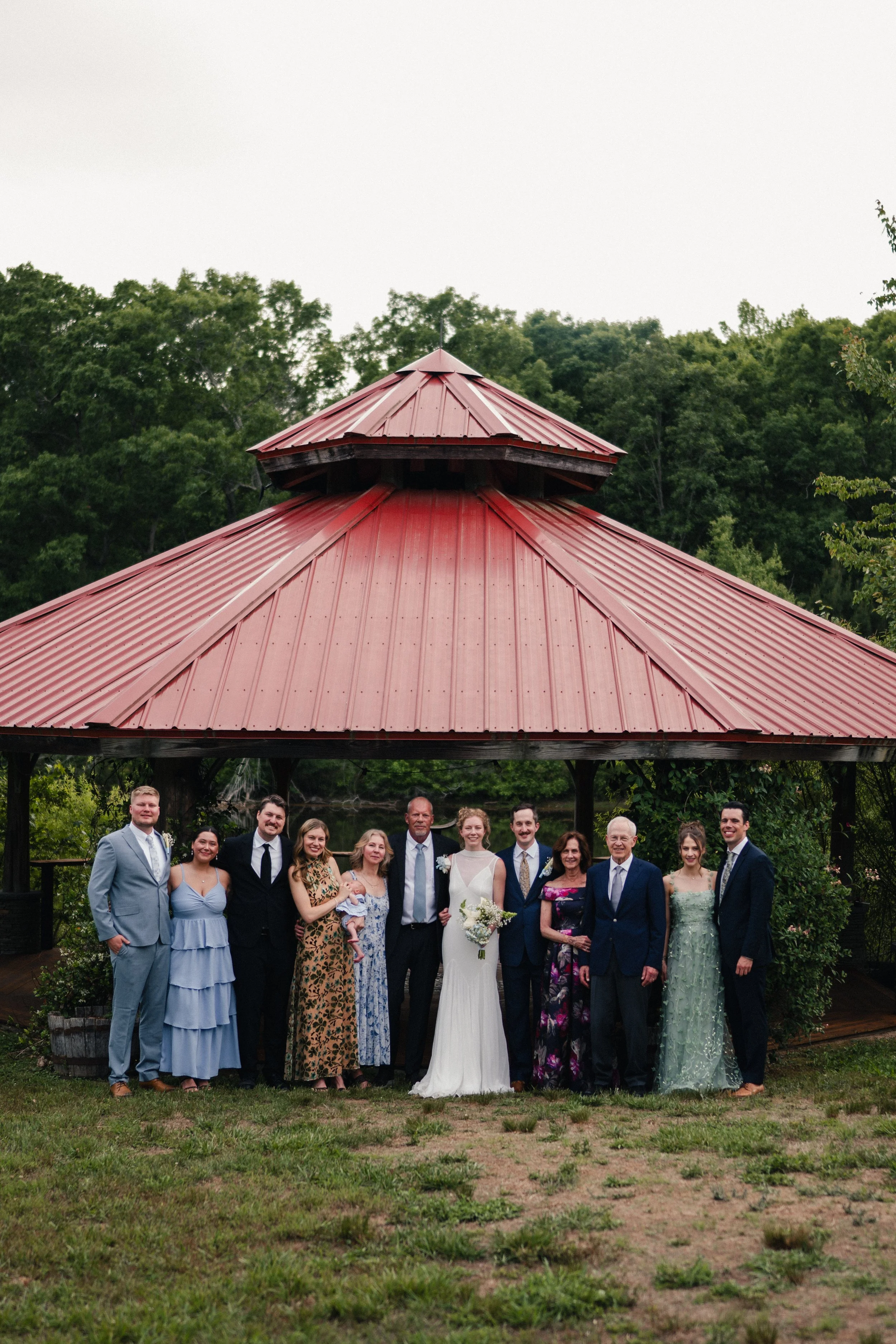 Group of people at a wedding posing outside under a red metal pavilion with a forest background.